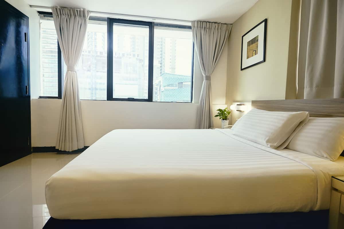 Een hotelkamer met een groot bed, een raam met gordijnen, een nachtkastje met een lamp en een lijst met een foto aan de muur.