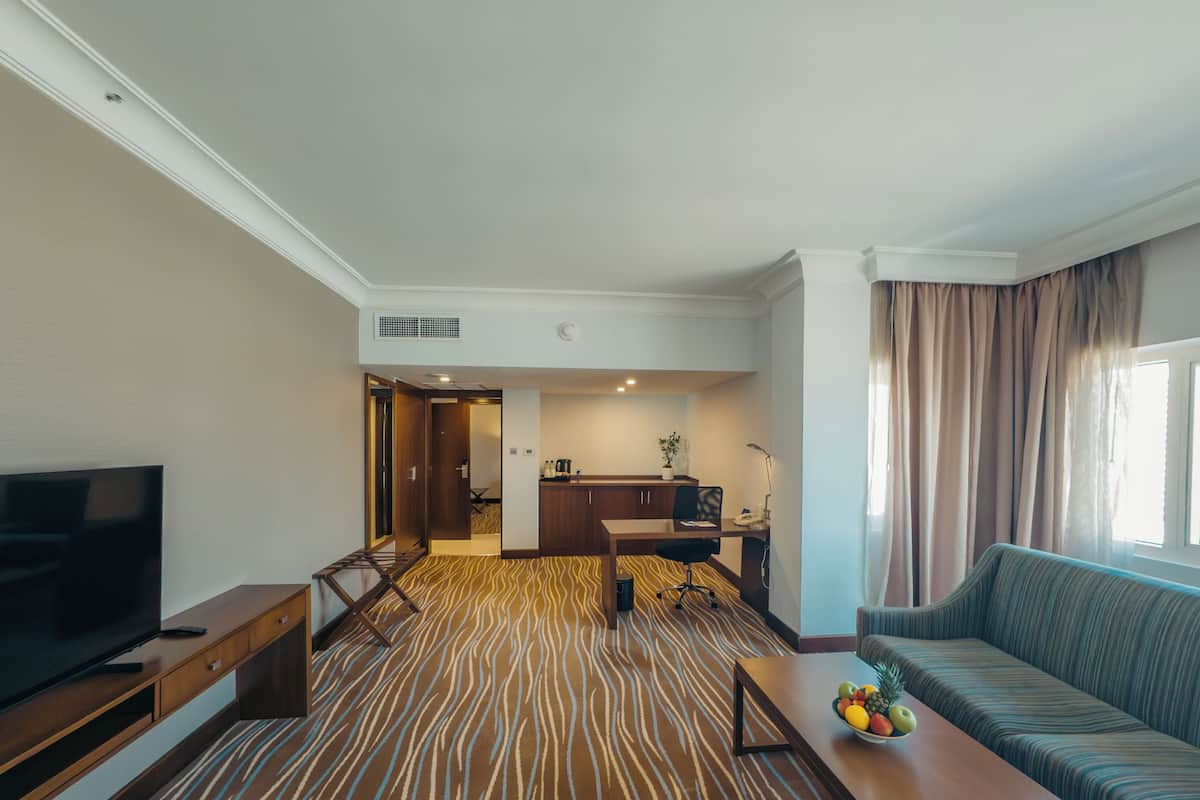 Habitación de hotel con televisión, sofá, mesa de centro con fruta, zona de cocina y alfombra estampada.