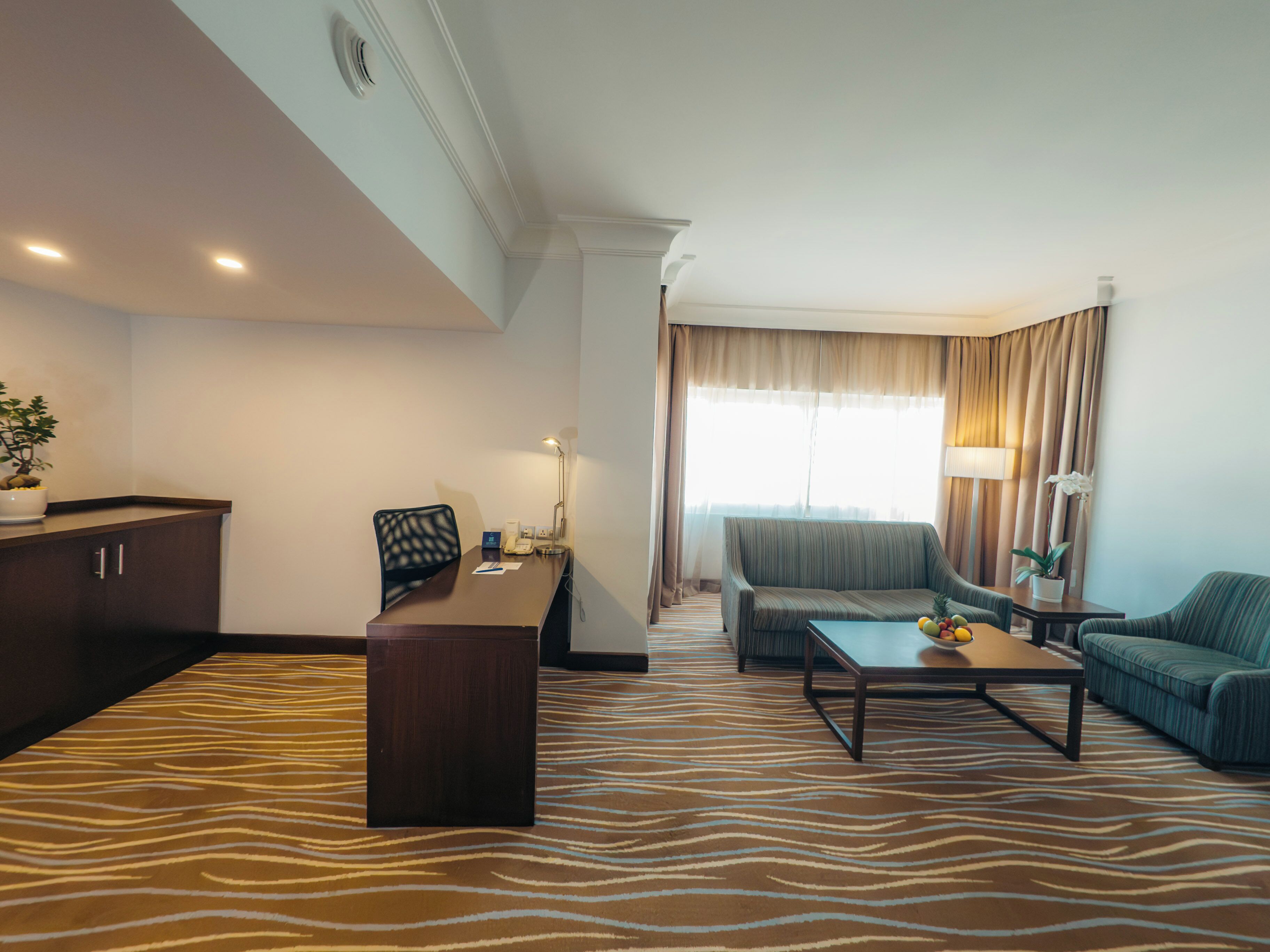 A modern hotel room with a desk, two chairs, a sofa, a coffee table, and a large window with curtains.