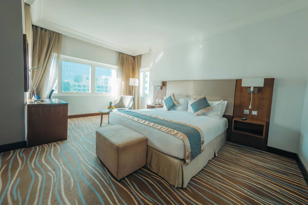 Una habitación de hotel con una cama grande, un escritorio, una silla, una mesita con frutero y vistas a la ciudad a través de la ventana.