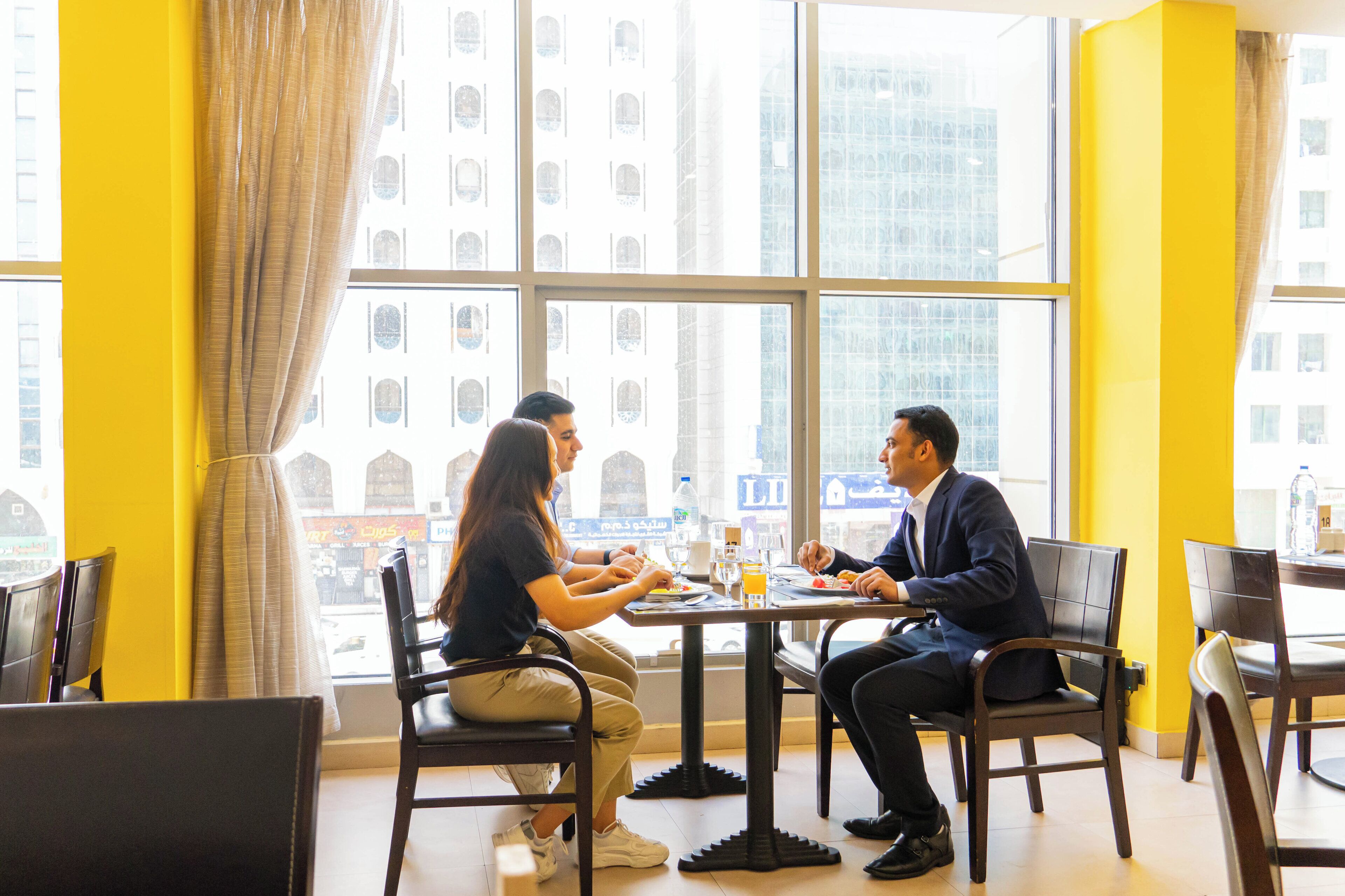 A dining area with a table and chairs, large windows with a view of buildings, and a yellow wall.