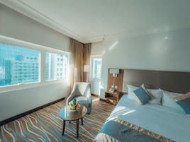Un cuarto de hotel con una cama grande, una mesita con fruta, una silla, una mesita de noche y vistas de la ciudad a través de amplias ventanas.