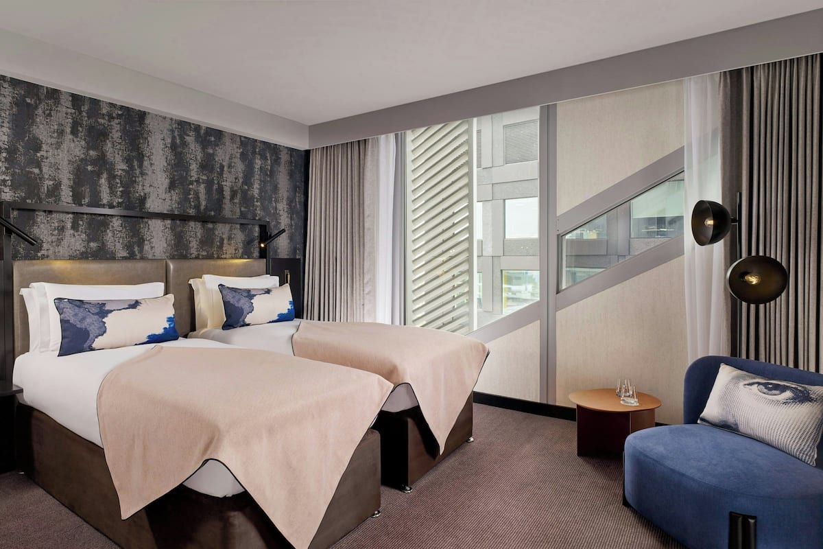 A hotel room with two beds, a sofa, a small table, and a large window with blinds.