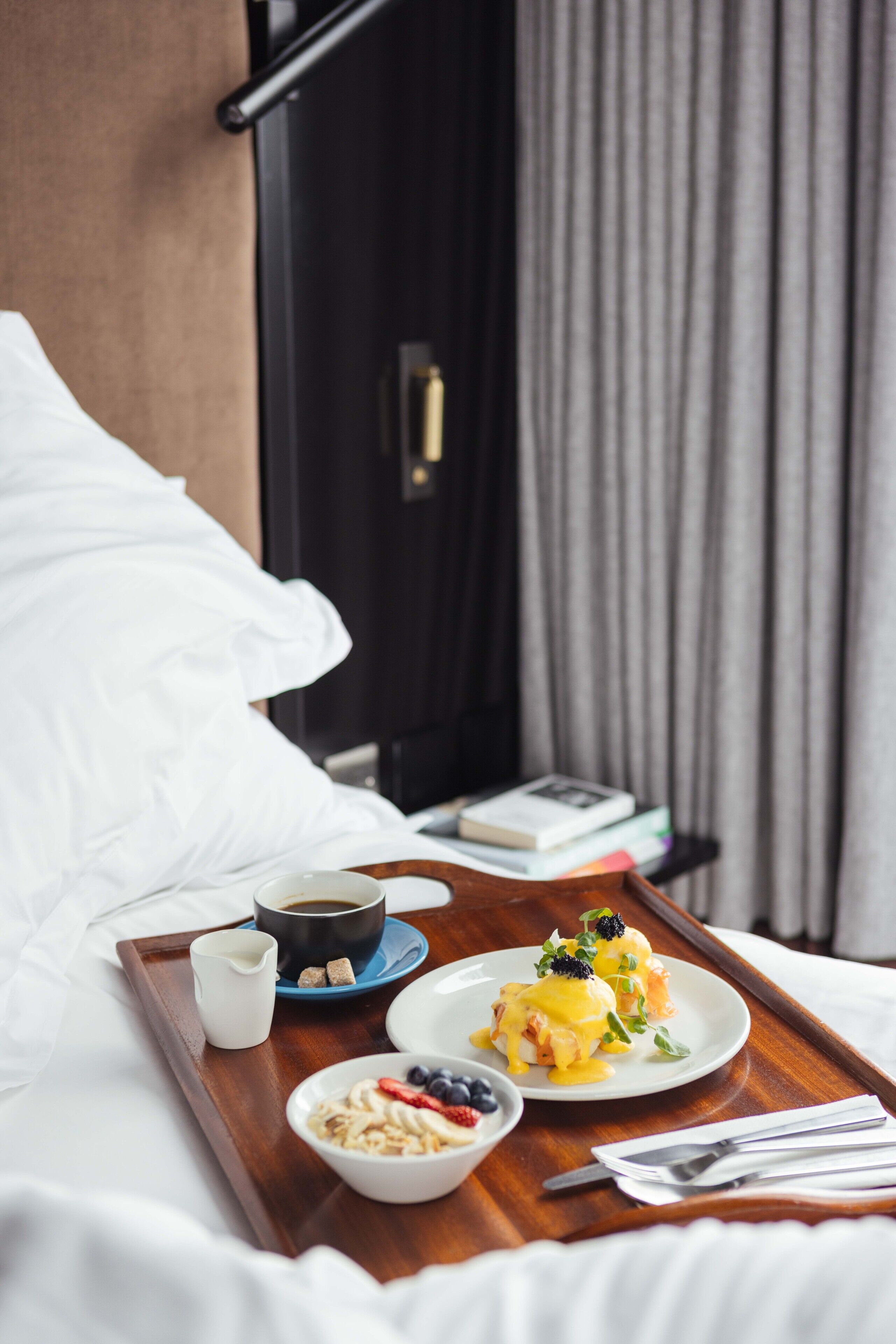 Bandeja de desayuno con huevos Benedict, café y bayas frescas sobre una cama de hotel.