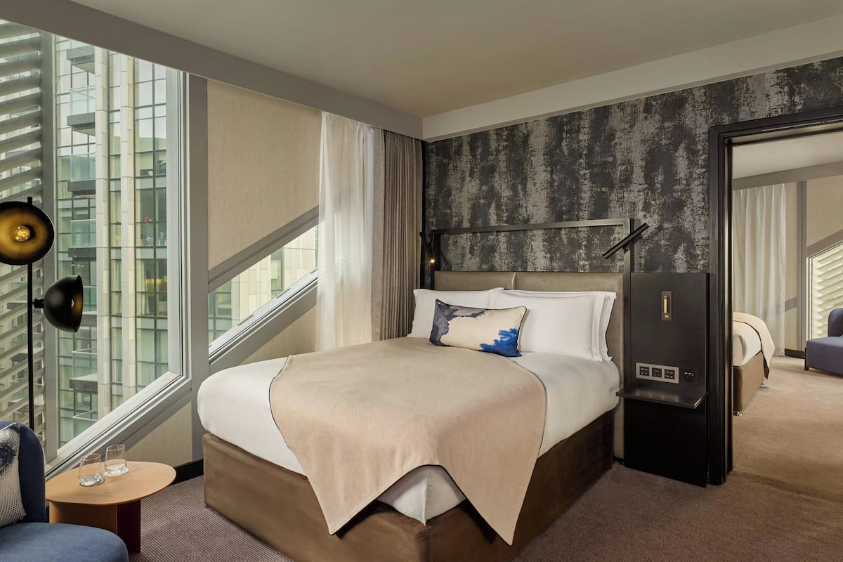 A modern hotel room with a large bed, a bedside table, a lamp, and a view of the city through large windows.