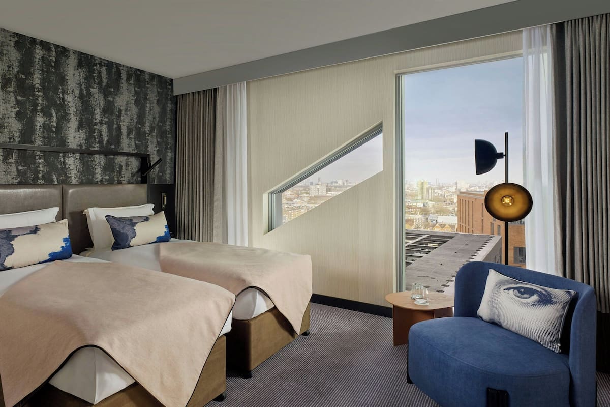 A hotel room with two beds, a city view, a desk with a lamp, and a blue armchair.