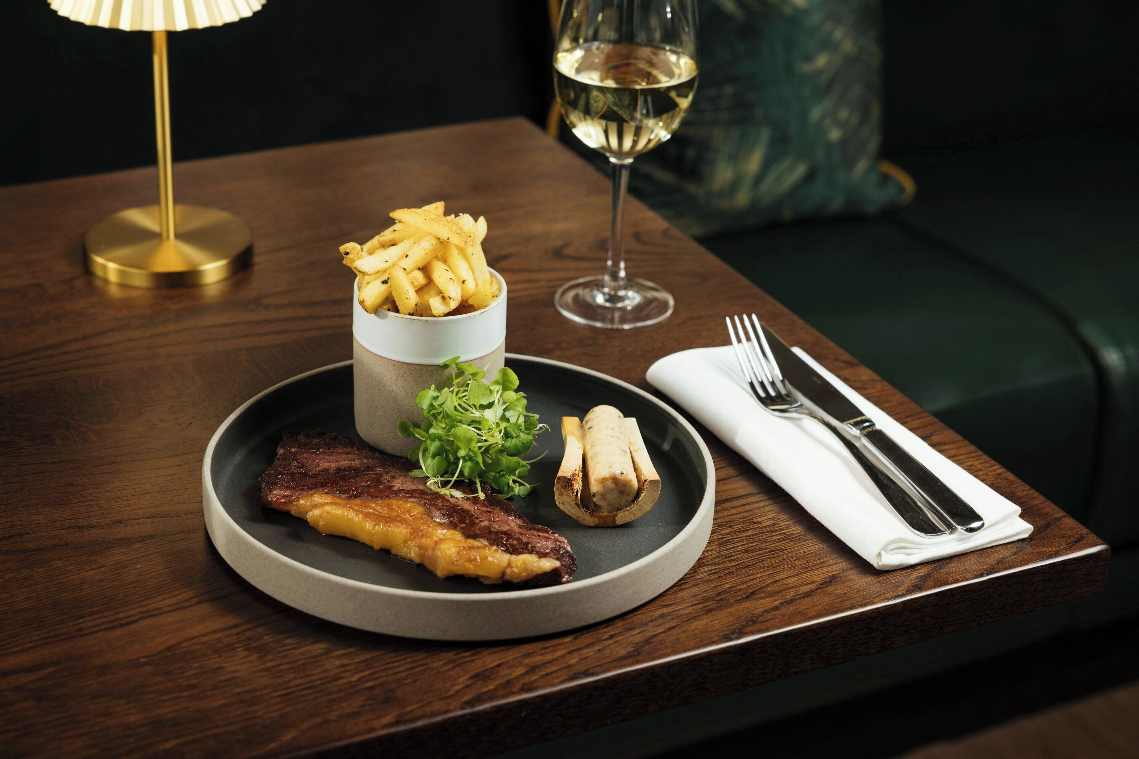 Ein Teller mit einem Steak, Pommes und Pilzen, begleitet von einem Glas Weißwein und einer Gabel und einem Messer auf einer Serviette, auf einem Holztisch platziert.
