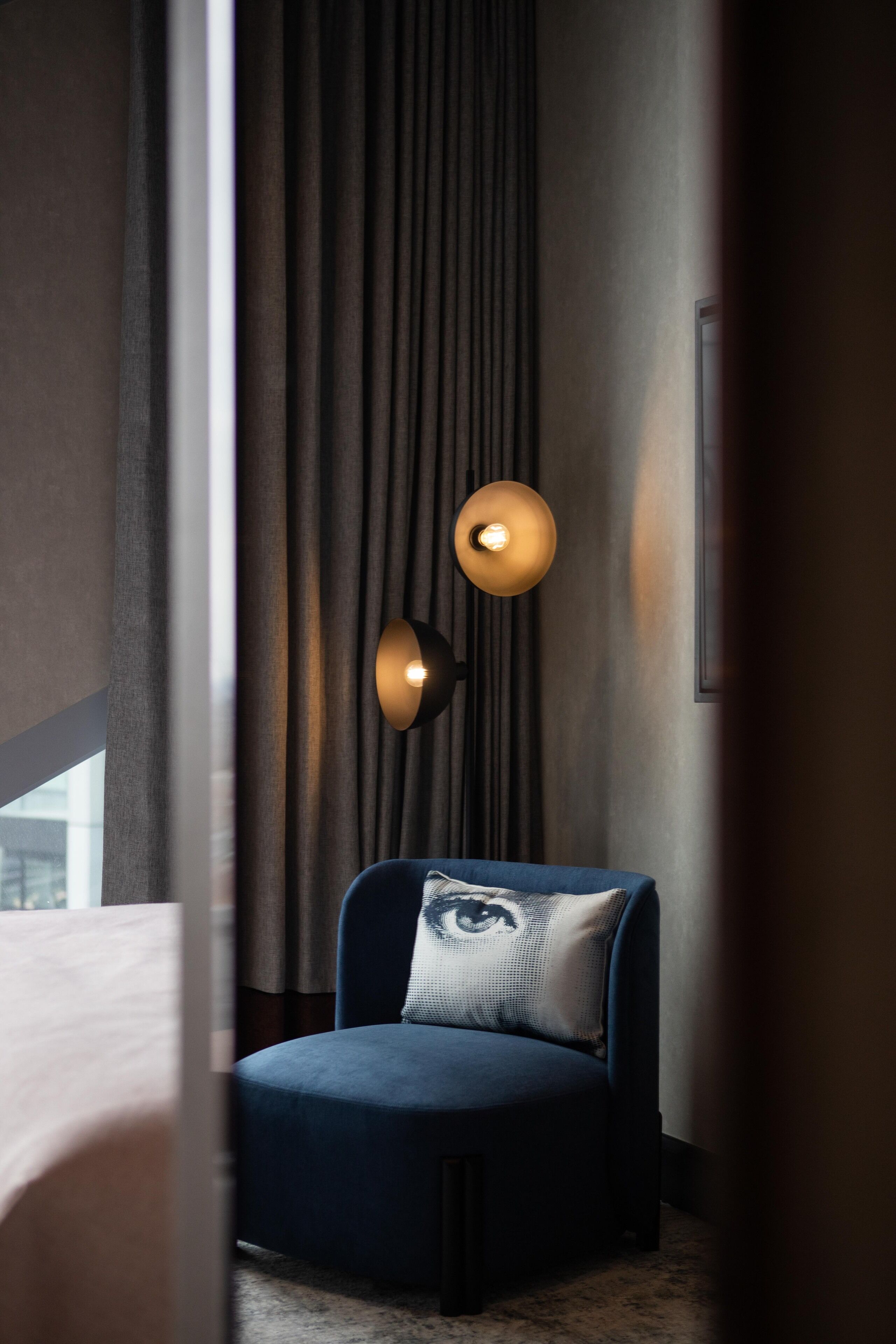 Ein modernes Hotelzimmer mit einem dunkelblauen Sessel, einem Zierkissen und einer Wandleuchte.