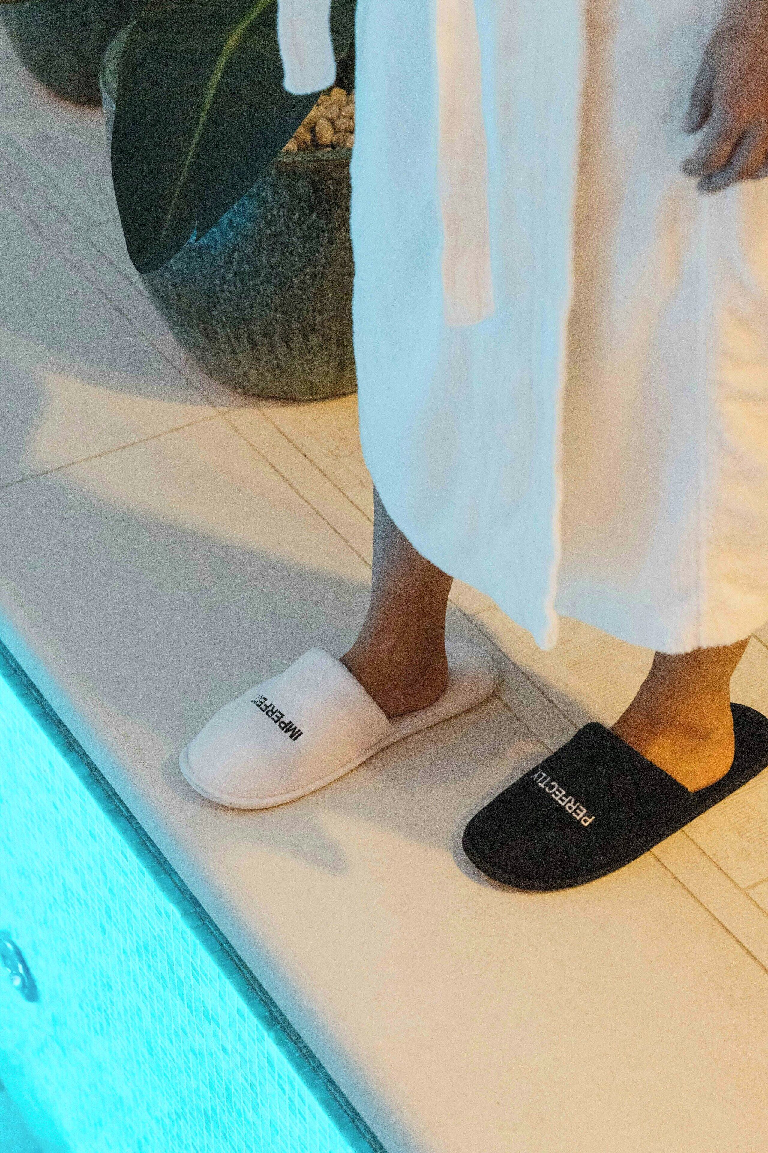 A person standing on a tiled floor wearing white slippers and black slippers.