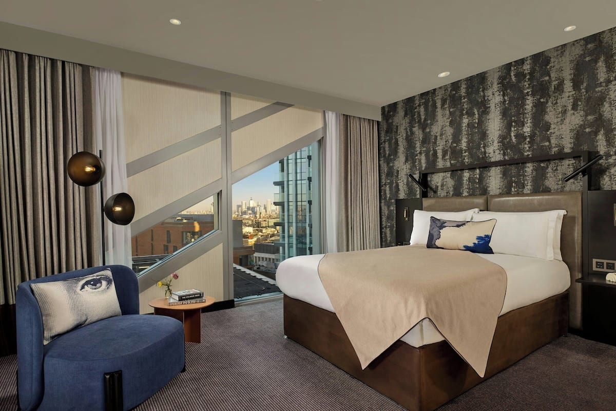 A hotel room with a large bed, a blue armchair, and a view of the city through floor-to-ceiling windows.