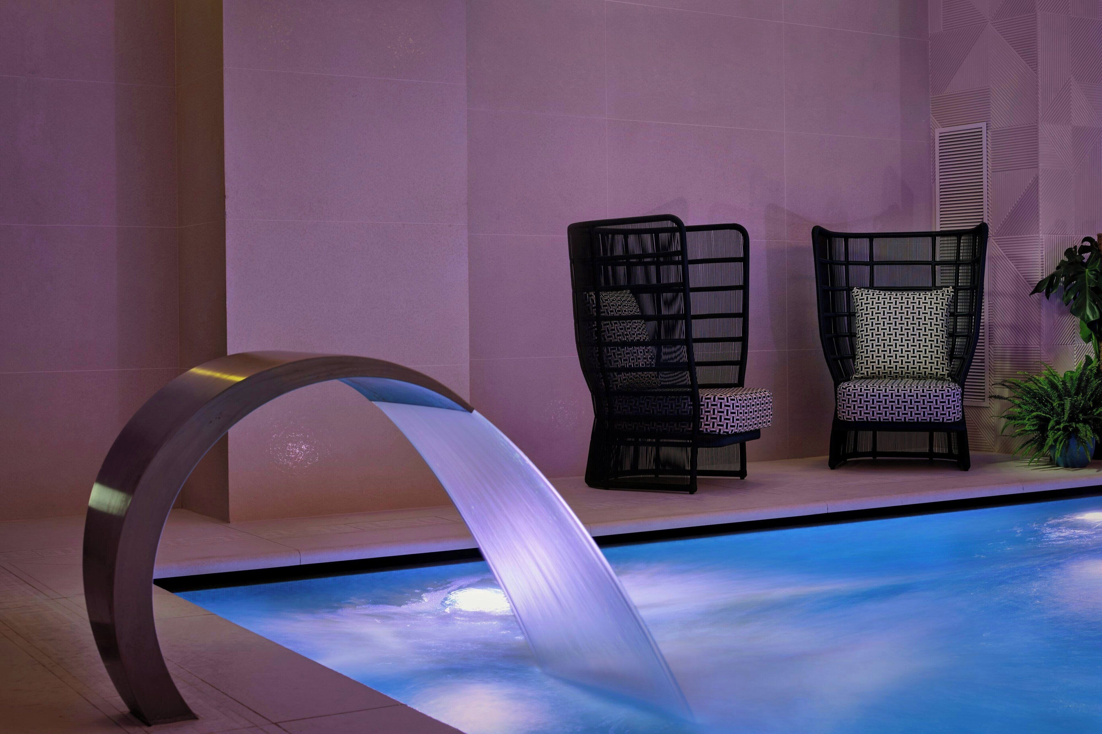 A modern indoor pool area with a unique metal arch structure, two wicker chairs with patterned cushions, and a potted plant.