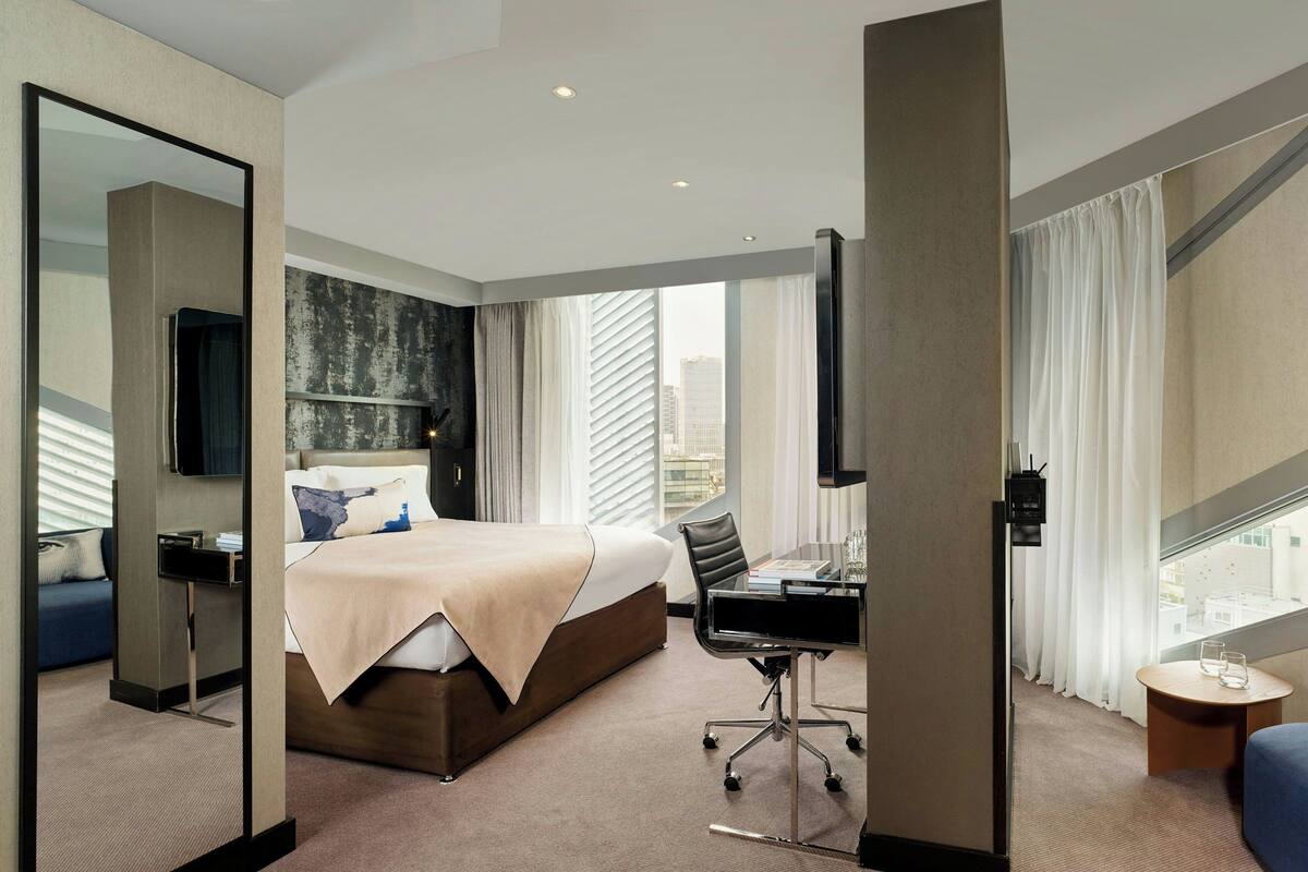 A modern hotel room with a large bed, a desk, and a view of the city through the window.