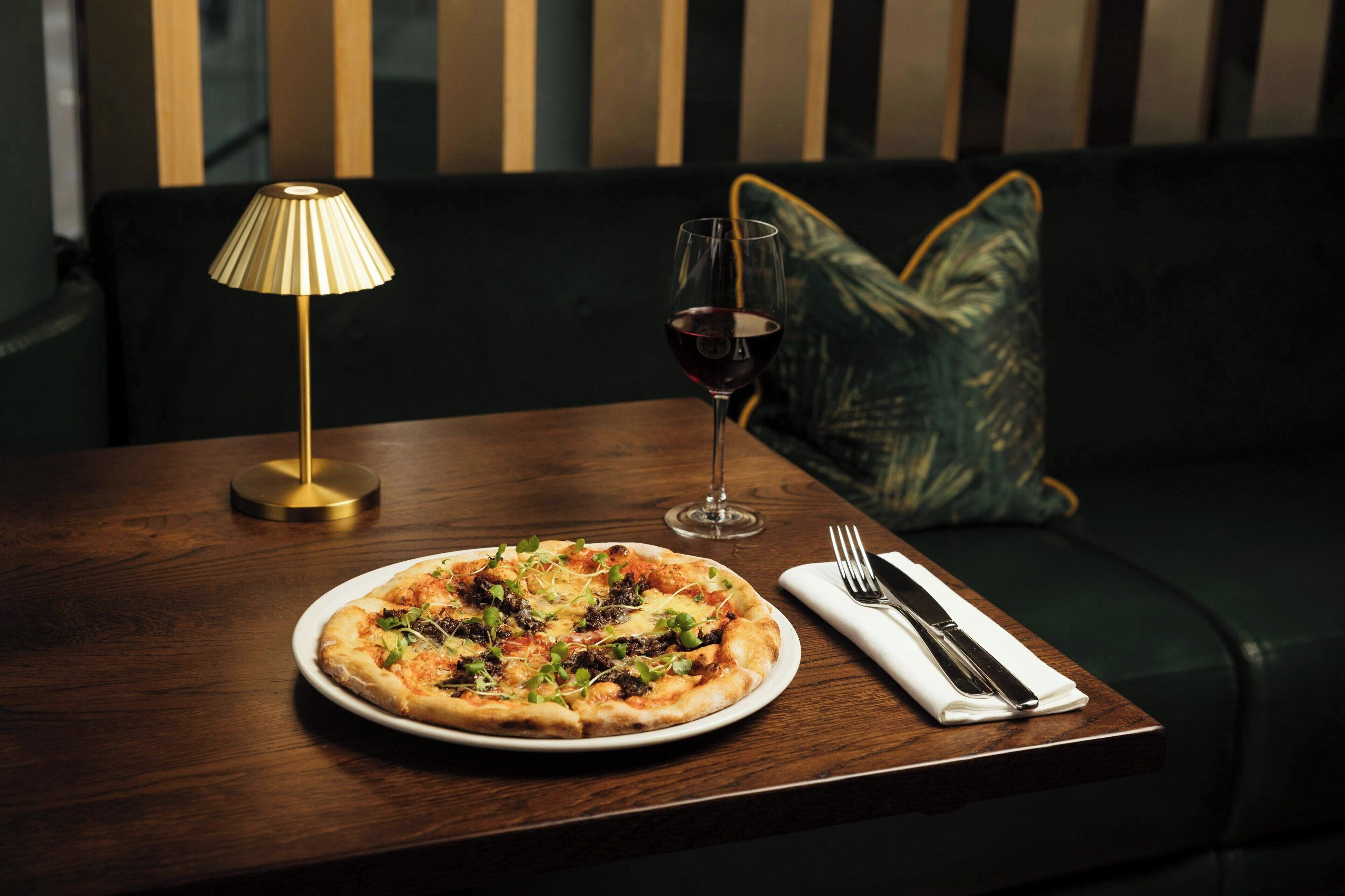 Pizza sobre una mesa de madera, con una lámpara y una copa de vino tinto sobre un sofá oscuro.