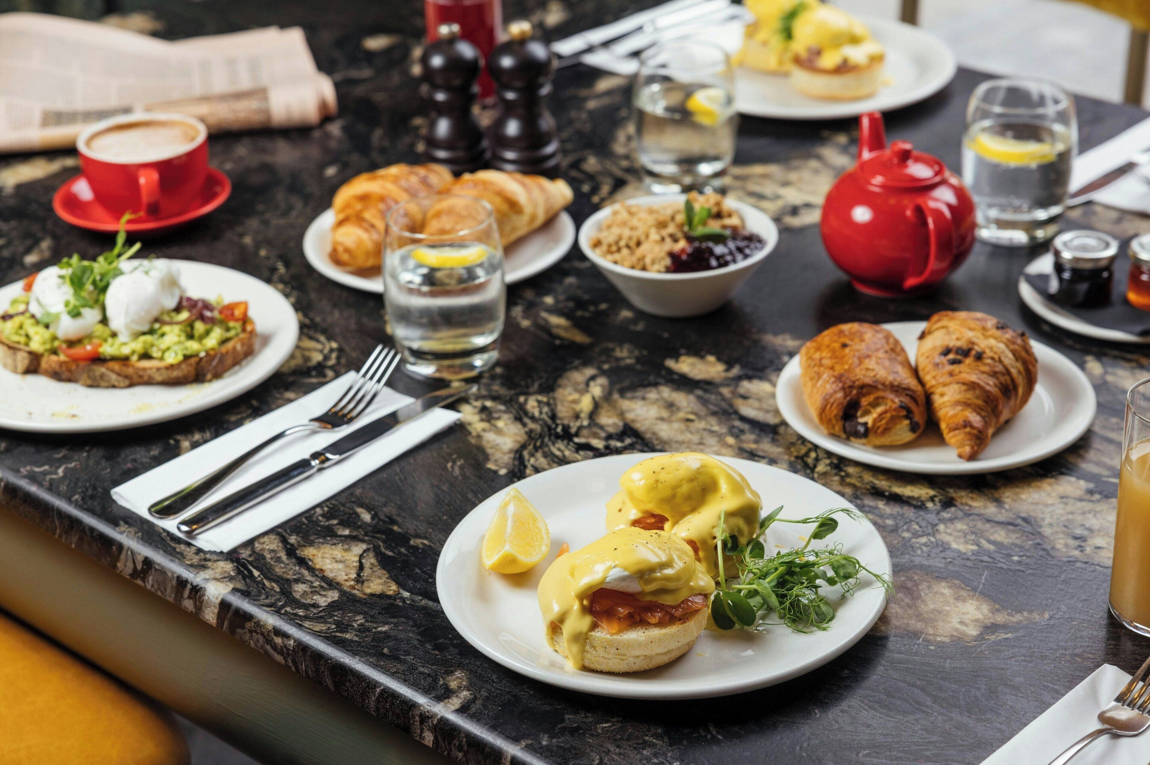 Une table dressée pour le petit-déjeuner, avec différents plats, notamment des œufs Bénédicte, des croissants et une tasse de café.