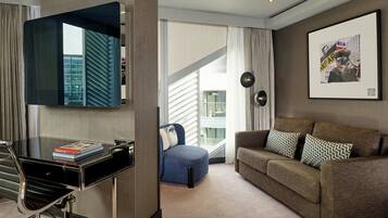 A modern hotel room with a glass desk, a blue armchair, a brown sofa, and a large window offering a view of a building.