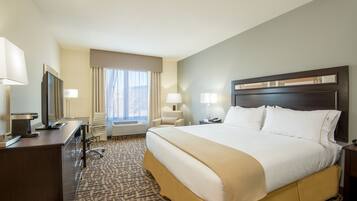 A hotel room with a large bed, a desk with a computer, a chair, a window with curtains, and a nightstand with a lamp.