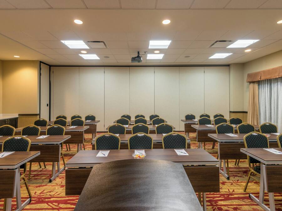 Un salón de conferencias con filas de mesas y sillas, una pantalla de proyector y un amplio ventanal con cortinas.