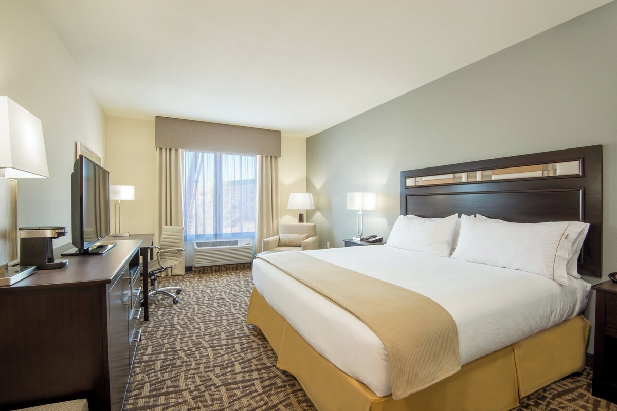 A hotel room with a large bed, a desk with a computer, a chair, a window with curtains, and a nightstand with a lamp.