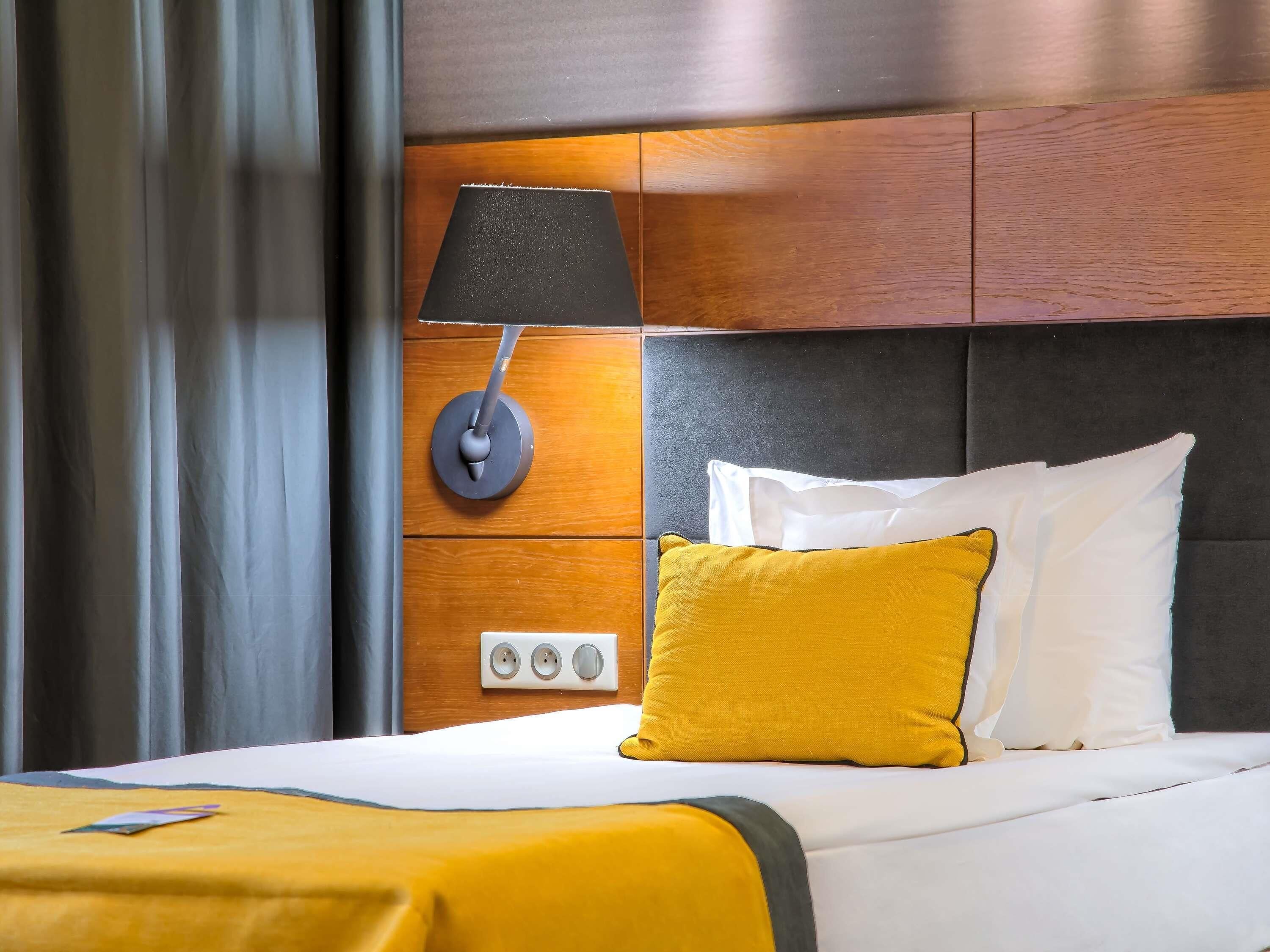 A hotel room with a bed, a wall-mounted lamp, a power outlet, and a window with curtains.