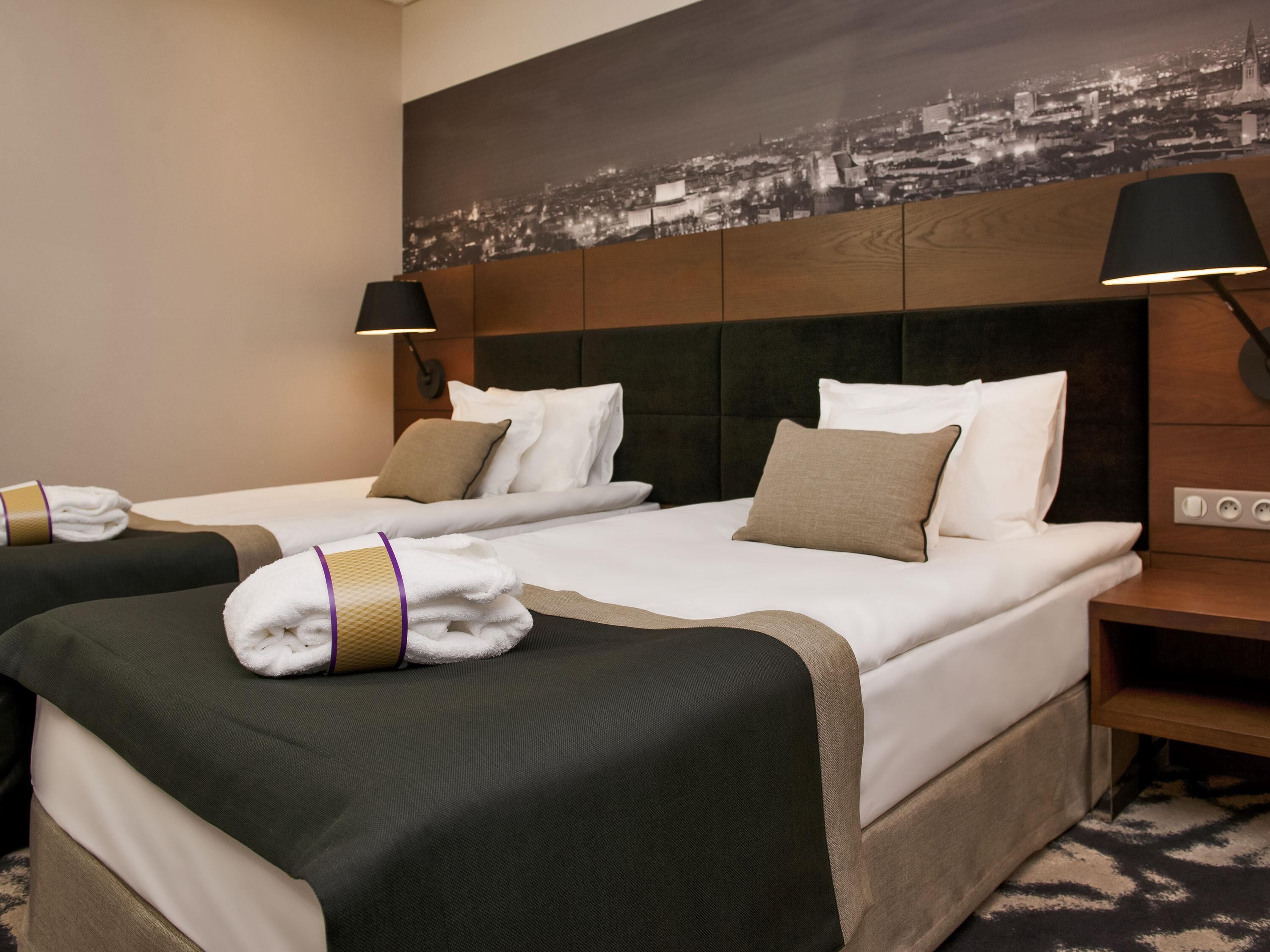 Habitación de hotel con dos camas, vista a la ciudad, cabecero de madera y mesita de noche con lámpara.