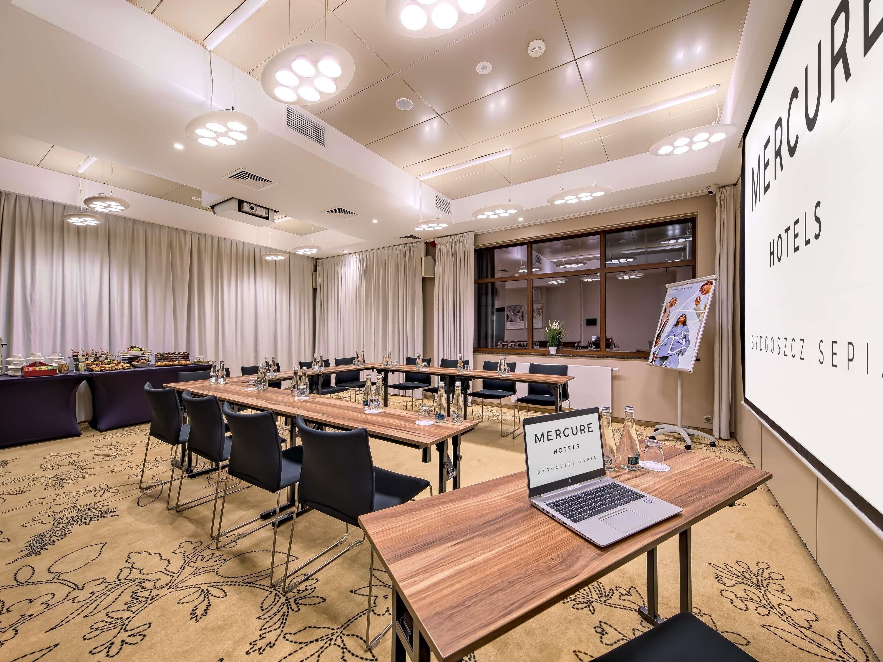 Un salón de conferencias con una mesa larga, sillas, una computadora portátil y una pantalla para presentaciones.