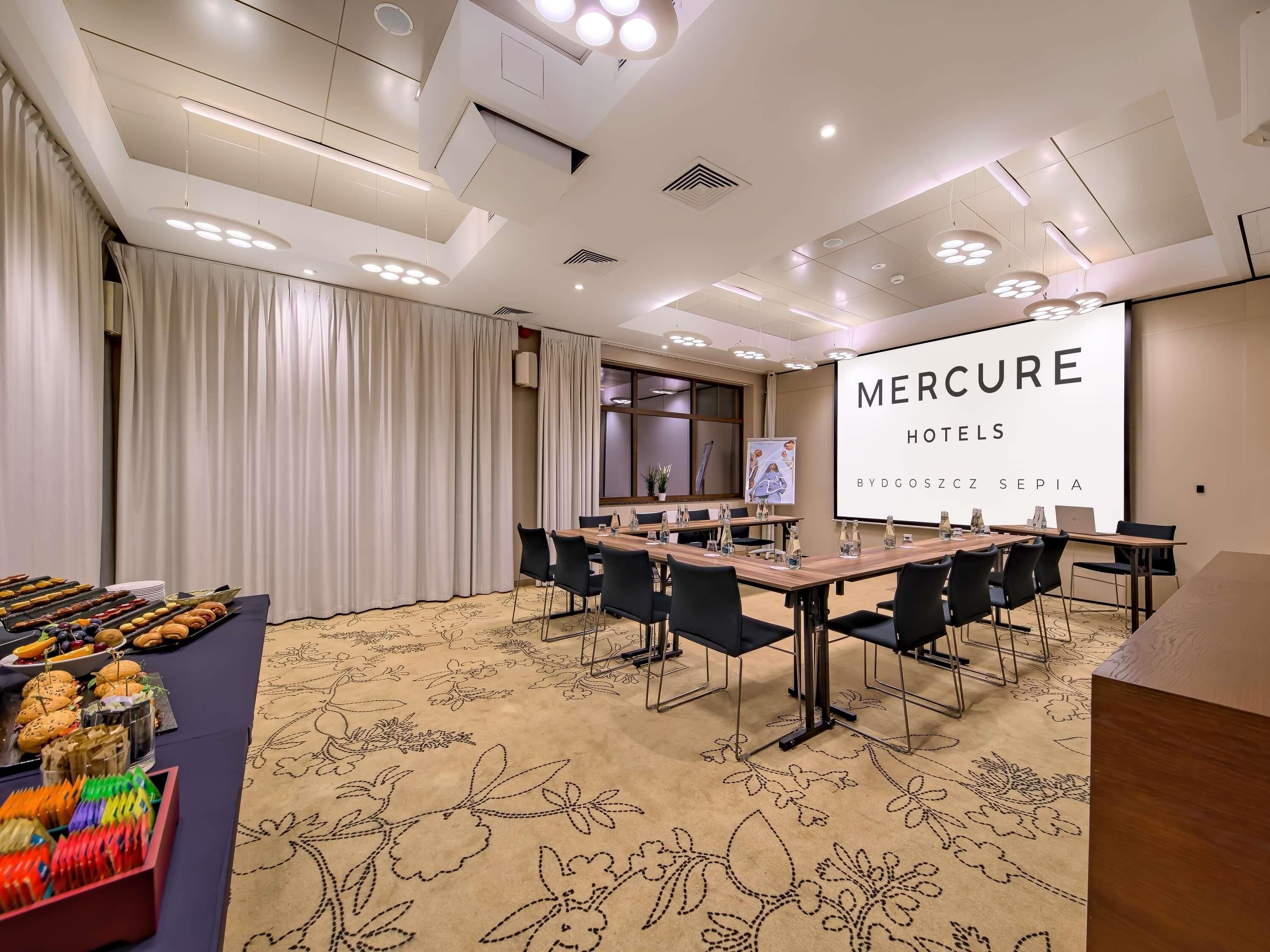 A conference room with a large screen displaying 'MERCURE HOTELS BYDGOSZCZ SEPIA', a long table with chairs, and a buffet table with snacks.