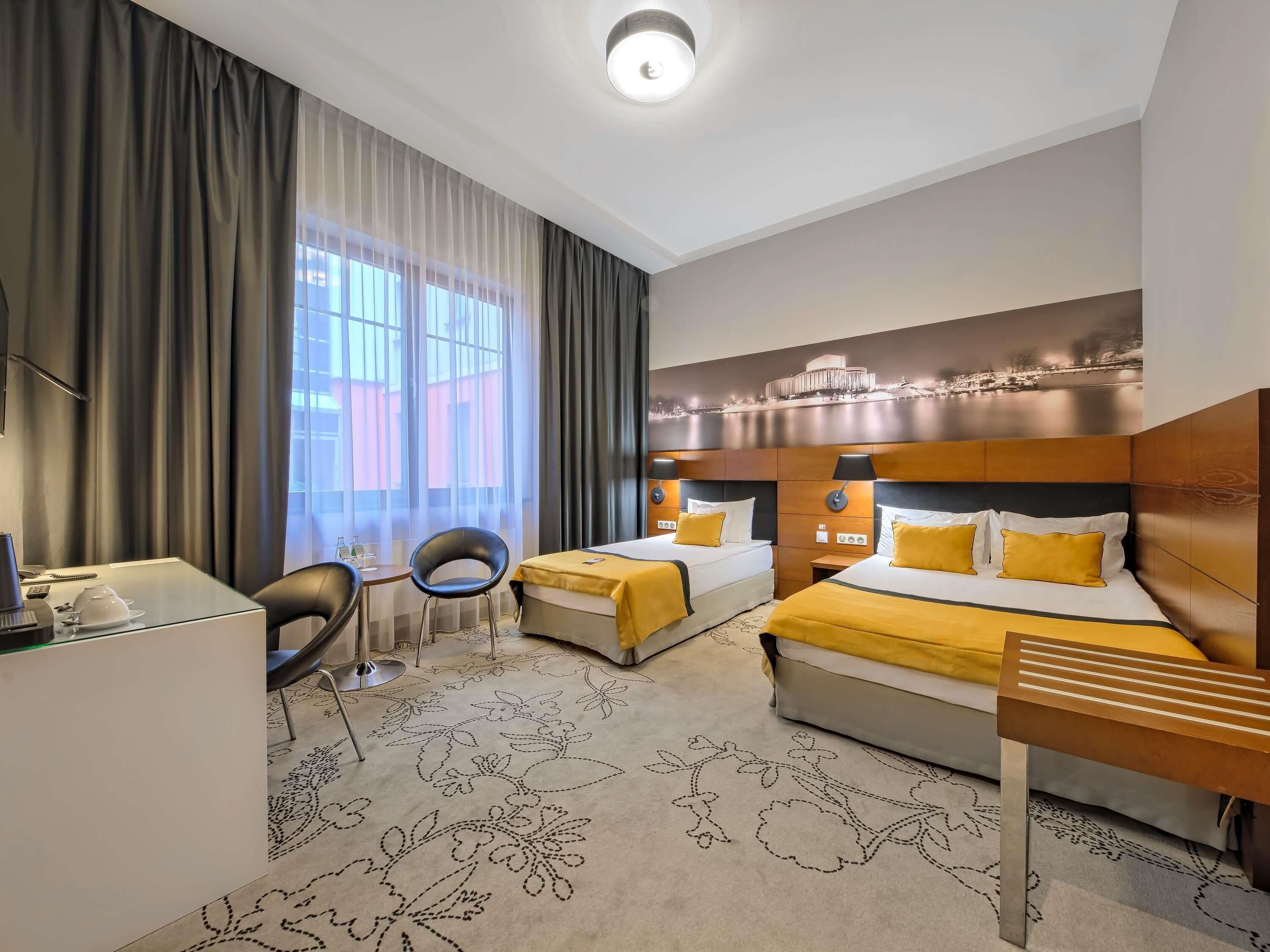 A hotel room with two beds, a desk, a chair, and a large window with curtains.