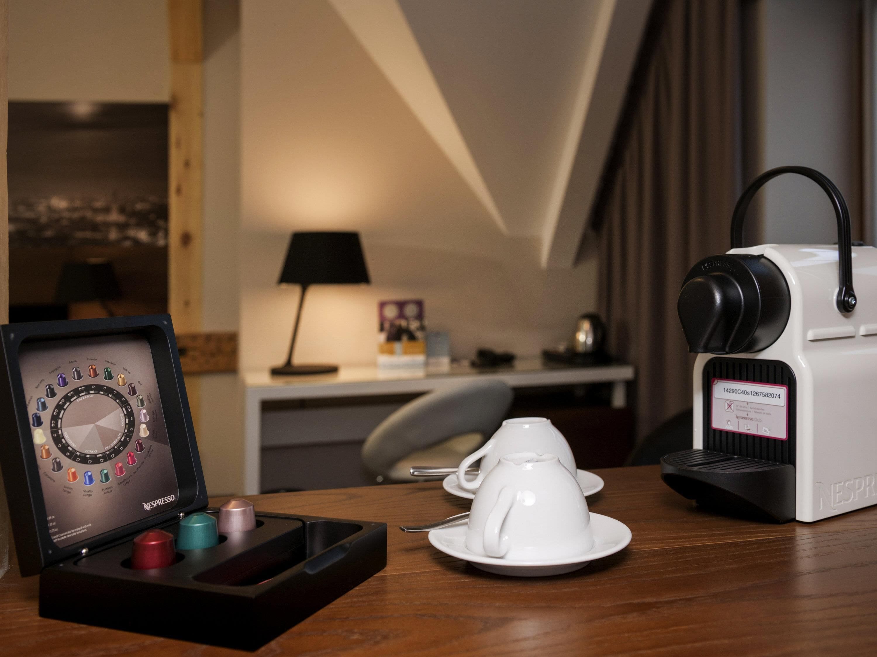 A coffee machine with a cup and saucer, a coffee pod holder, and a lamp on a table in a hotel room.