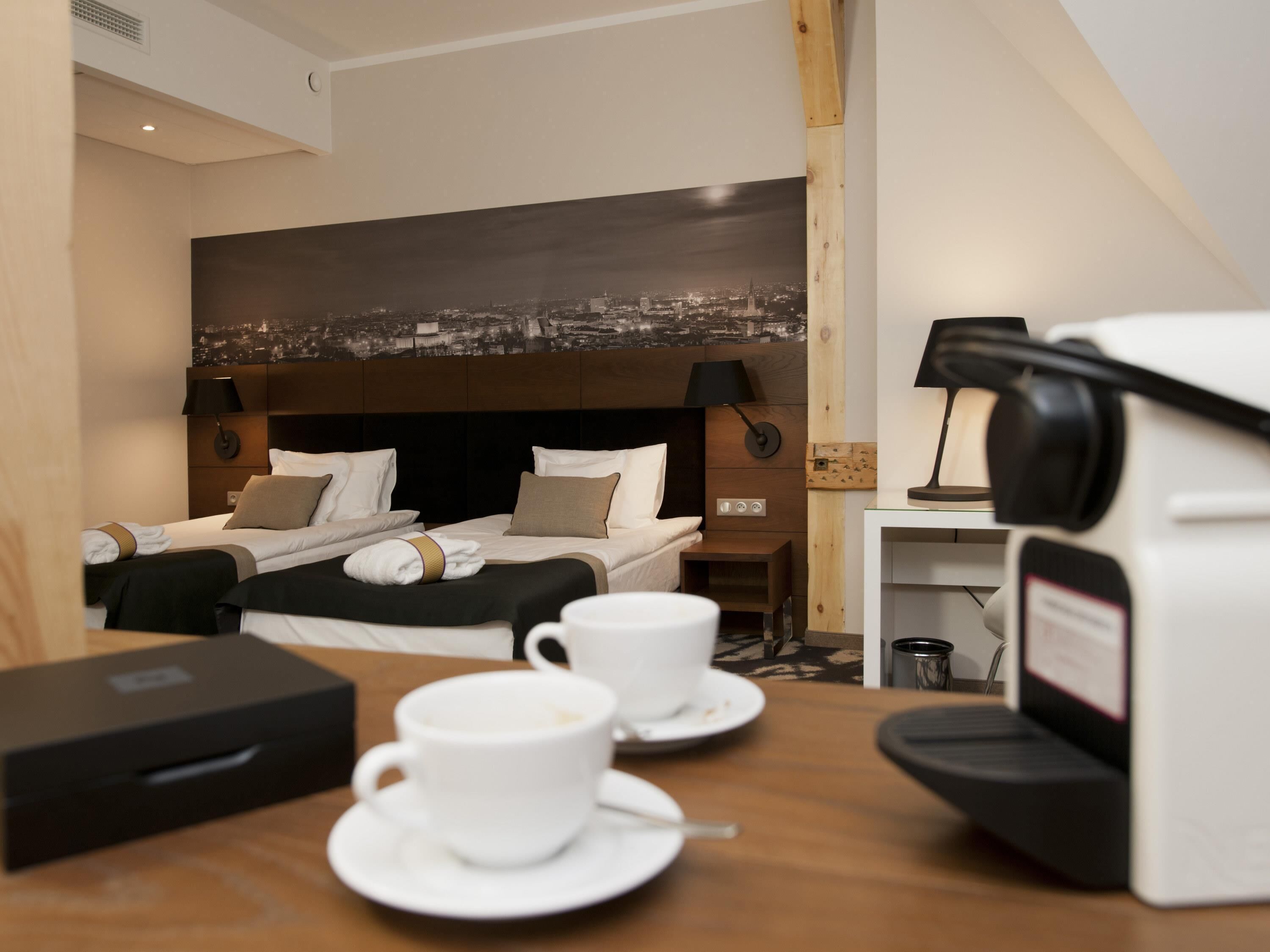 A hotel room with two beds, a cityscape view, a coffee machine, and a table with a cup and saucer.
