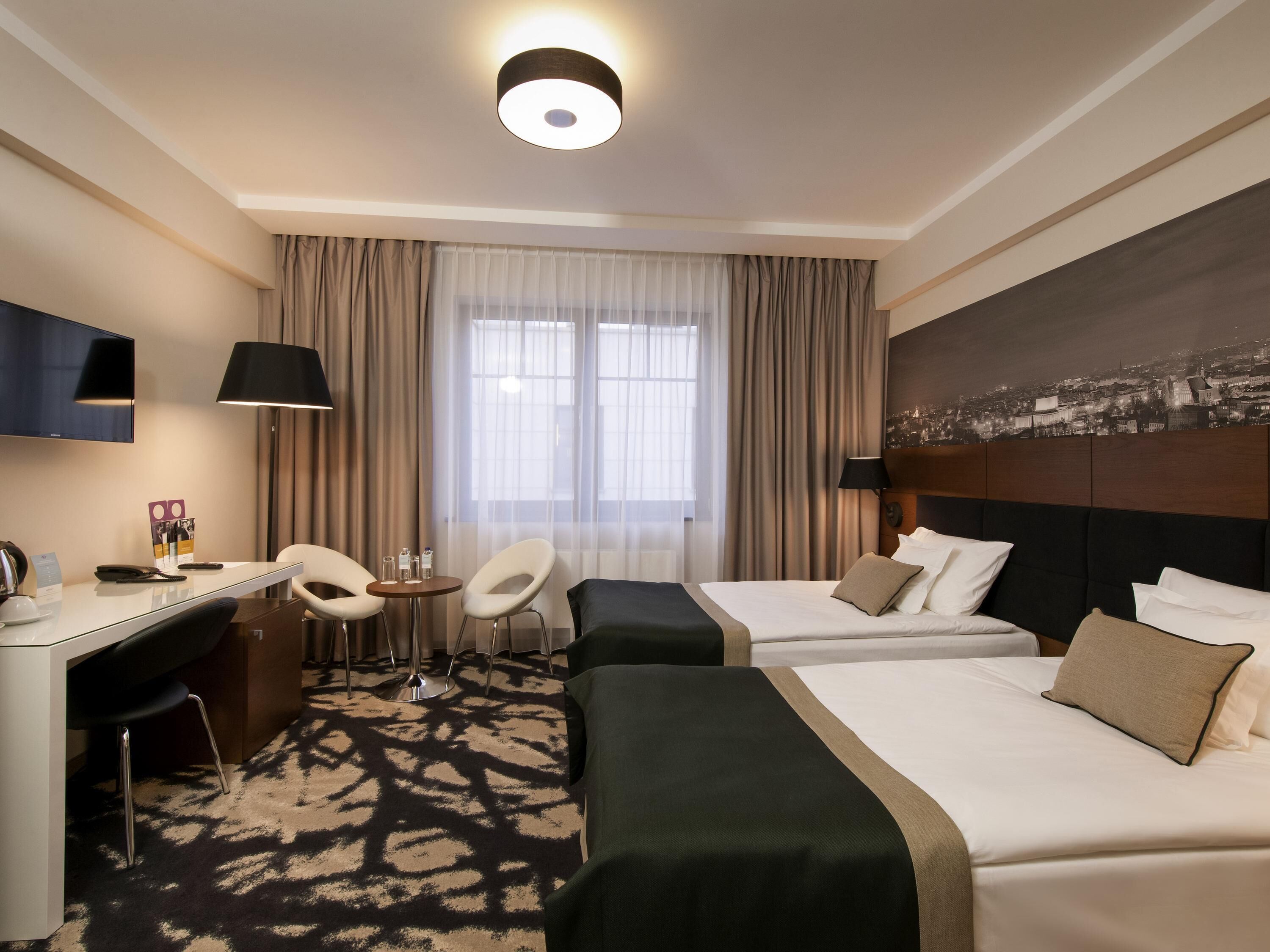 A hotel room with two beds, a desk, chairs, a TV, and a cityscape view.