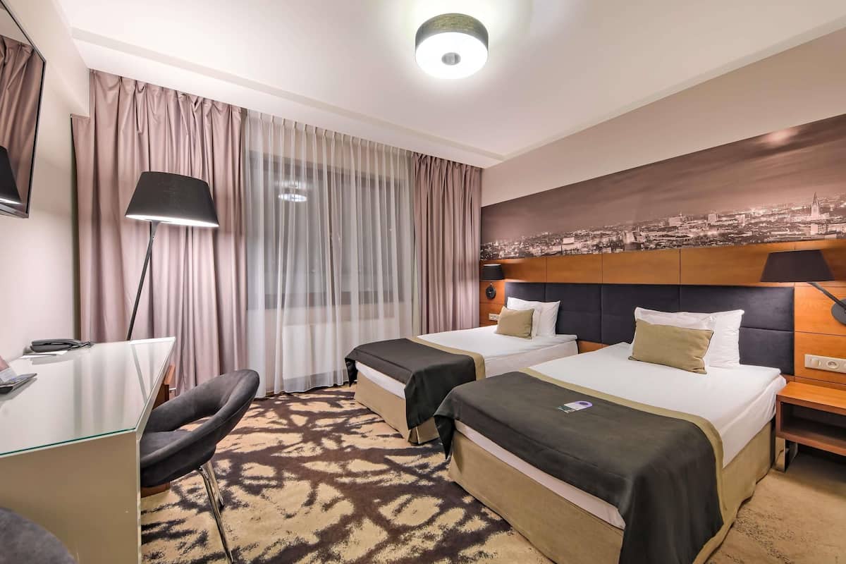 Ein Hotelzimmer mit zwei Betten, einem Schreibtisch, einem Stuhl, einem Stadtbild-Wandgemälde und einem gemusterten Teppich.