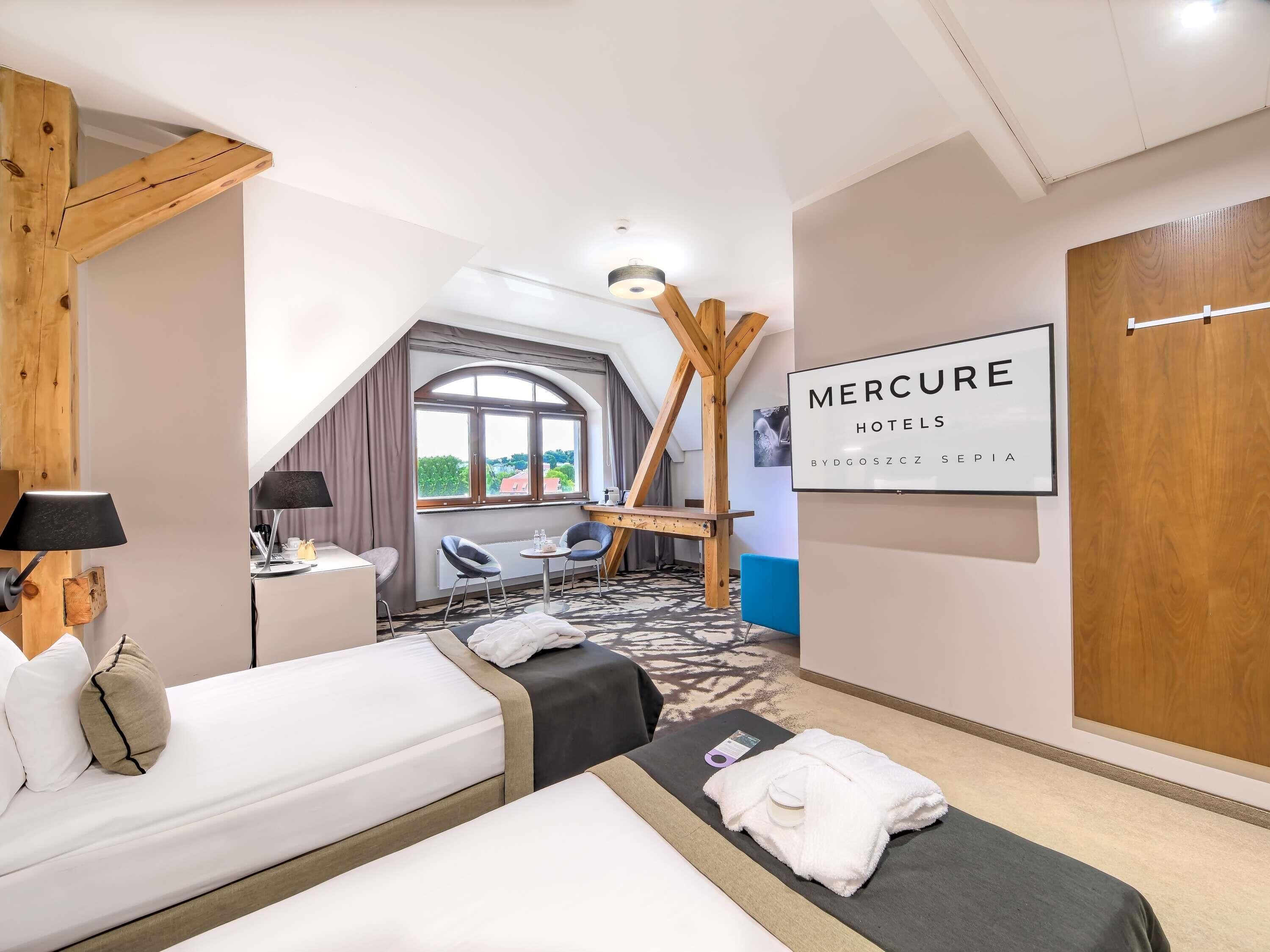 A hotel room with two beds, a desk, a chair, a window with a view, and a sign for Mercure Hotels.