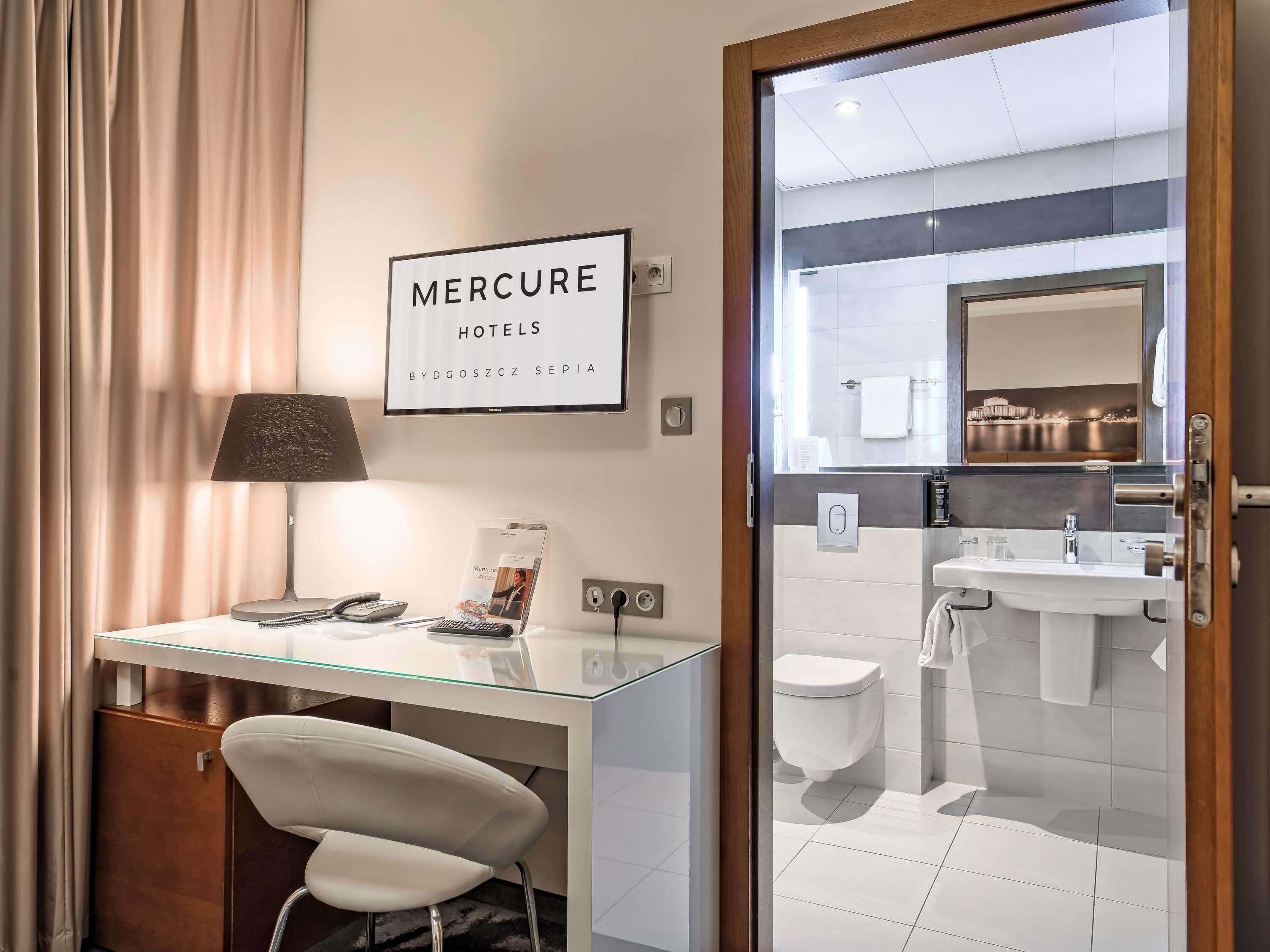 Une salle de bain d’hôtel moderne avec un bureau en verre, une chaise, une lampe, un téléphone et un panneau encadré placé au-dessus du bureau.