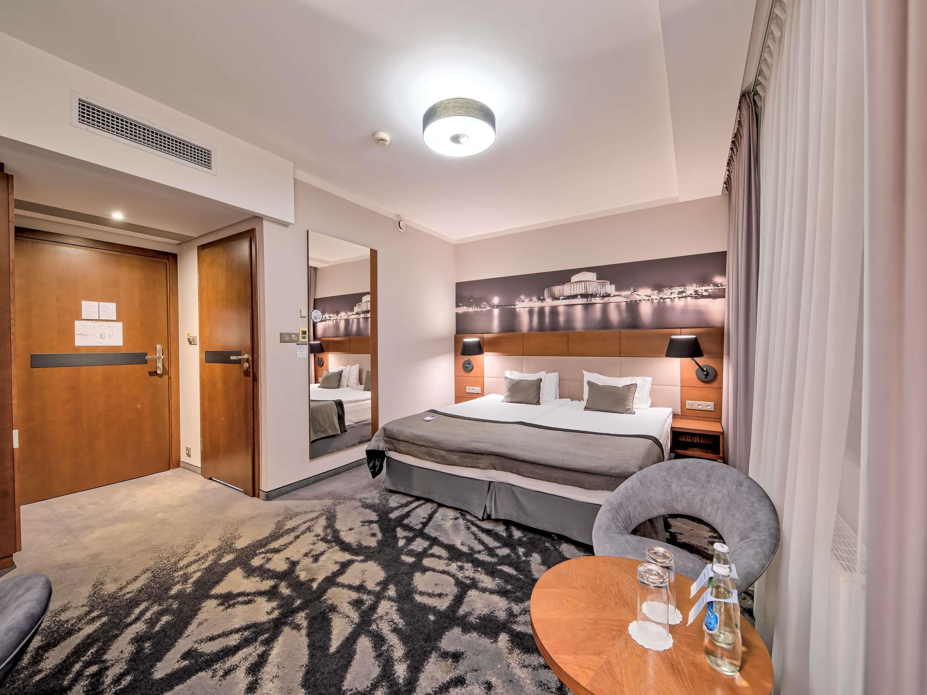Ett hotellrum med en säng, sängbord, en stol, ett runt bord, en tavla på väggen och en dörr.