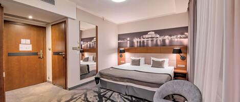 Habitación de hotel con cama, mesitas de noche, una silla, una mesa redonda, un cuadro en la pared y una puerta.