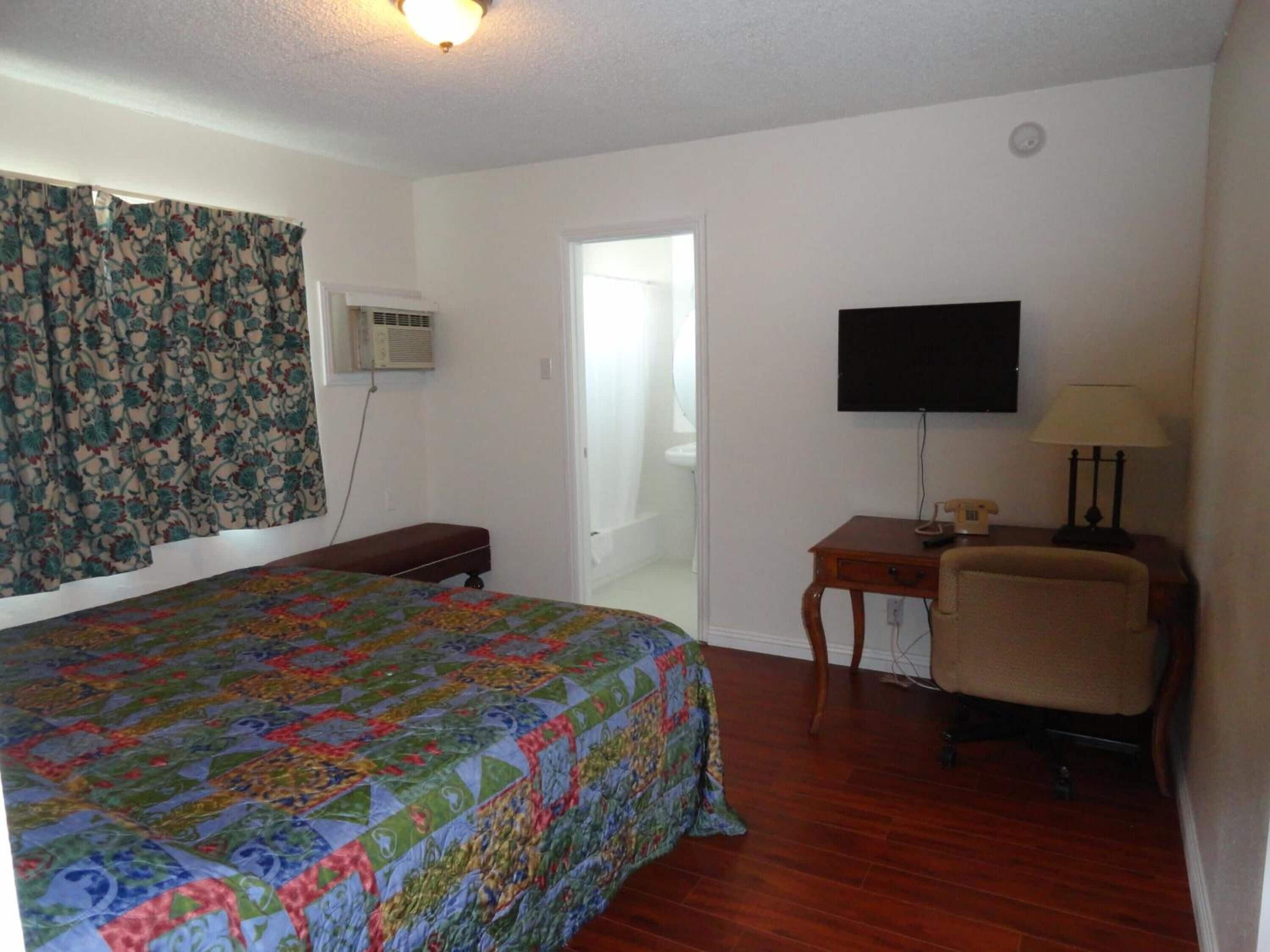 A bedroom with a bed, a desk, a chair, a television, and an air conditioning unit.