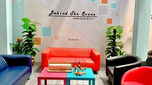 A room with a red sofa, a blue sofa, a black chair, and a small table with a toy. There are two potted plants and a wall with a sign that reads "Behind The Scene Hotel Club @ Samui".