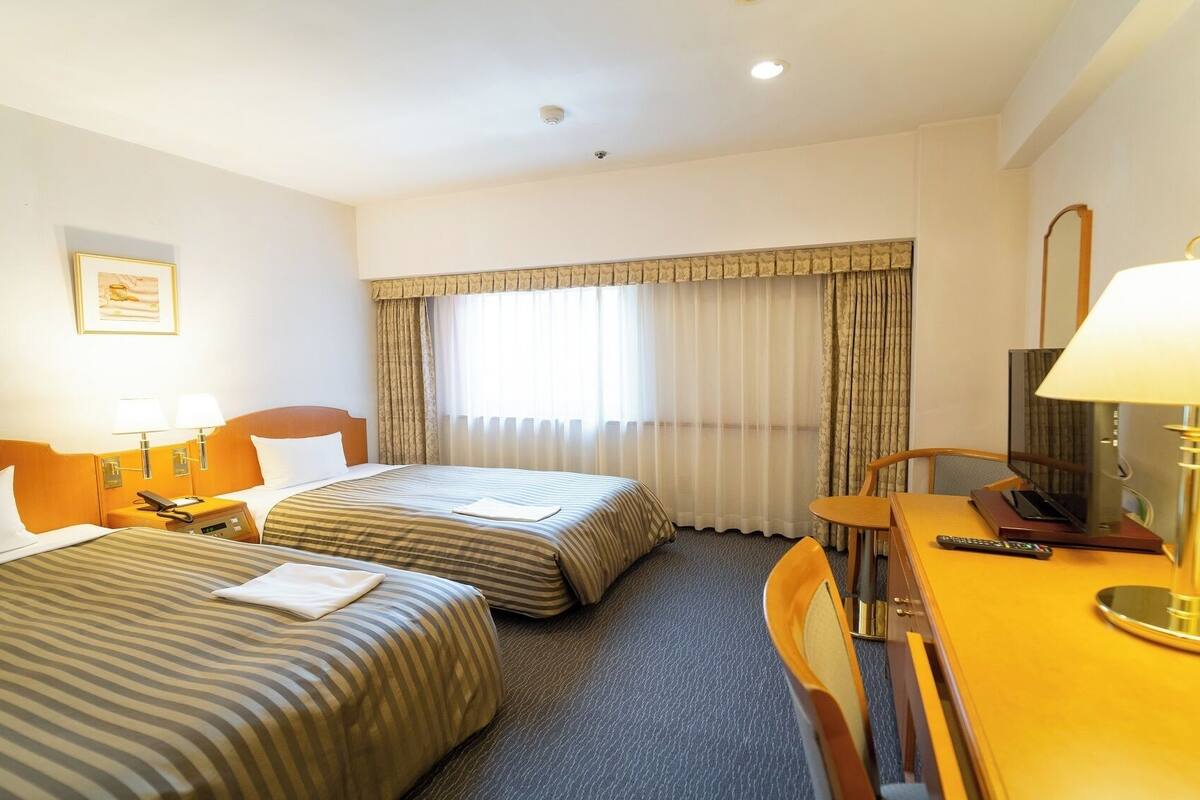 Habitación de hotel con dos camas, un escritorio con televisor y una ventana con cortinas.