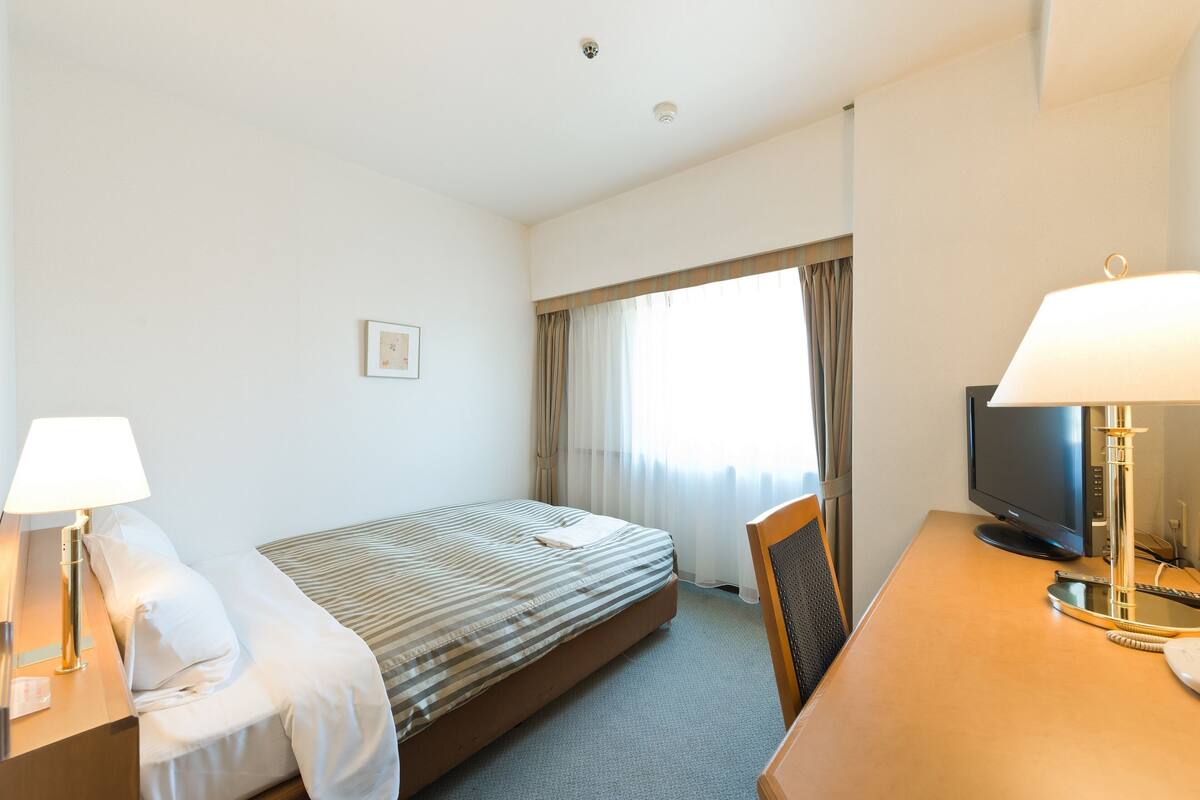 Habitación de hotel con cama, escritorio con lámpara, televisión y ventana con cortinas.