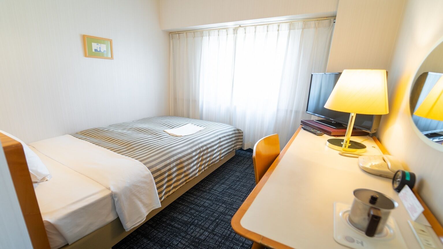 Une chambre d’hôtel avec un lit simple, un bureau avec une lampe, une télévision et une fenêtre avec des rideaux.