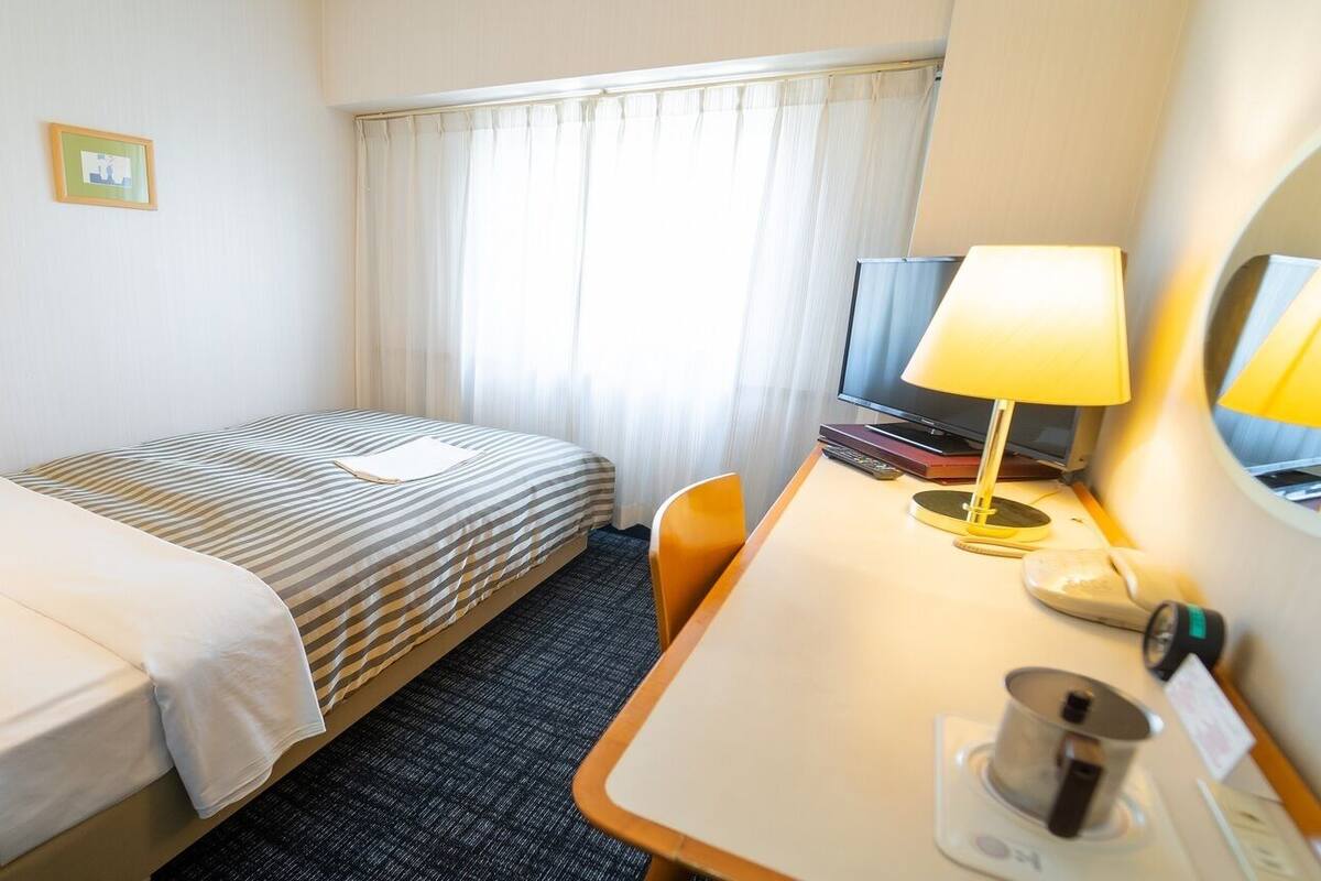 Habitación de hotel con una cama individual, un escritorio con lámpara, un televisor y una ventana con cortinas.