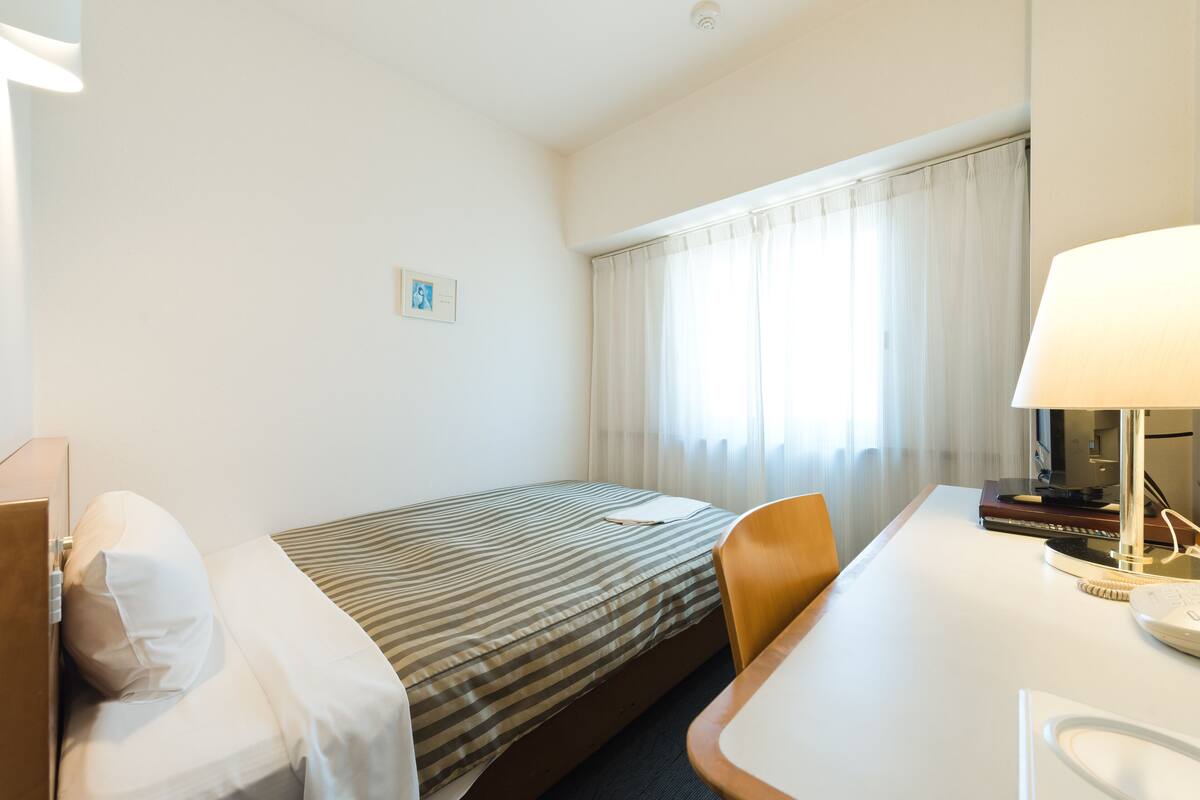 Habitación de hotel con una cama individual, un escritorio con silla, una lámpara y una ventana con cortinas.
