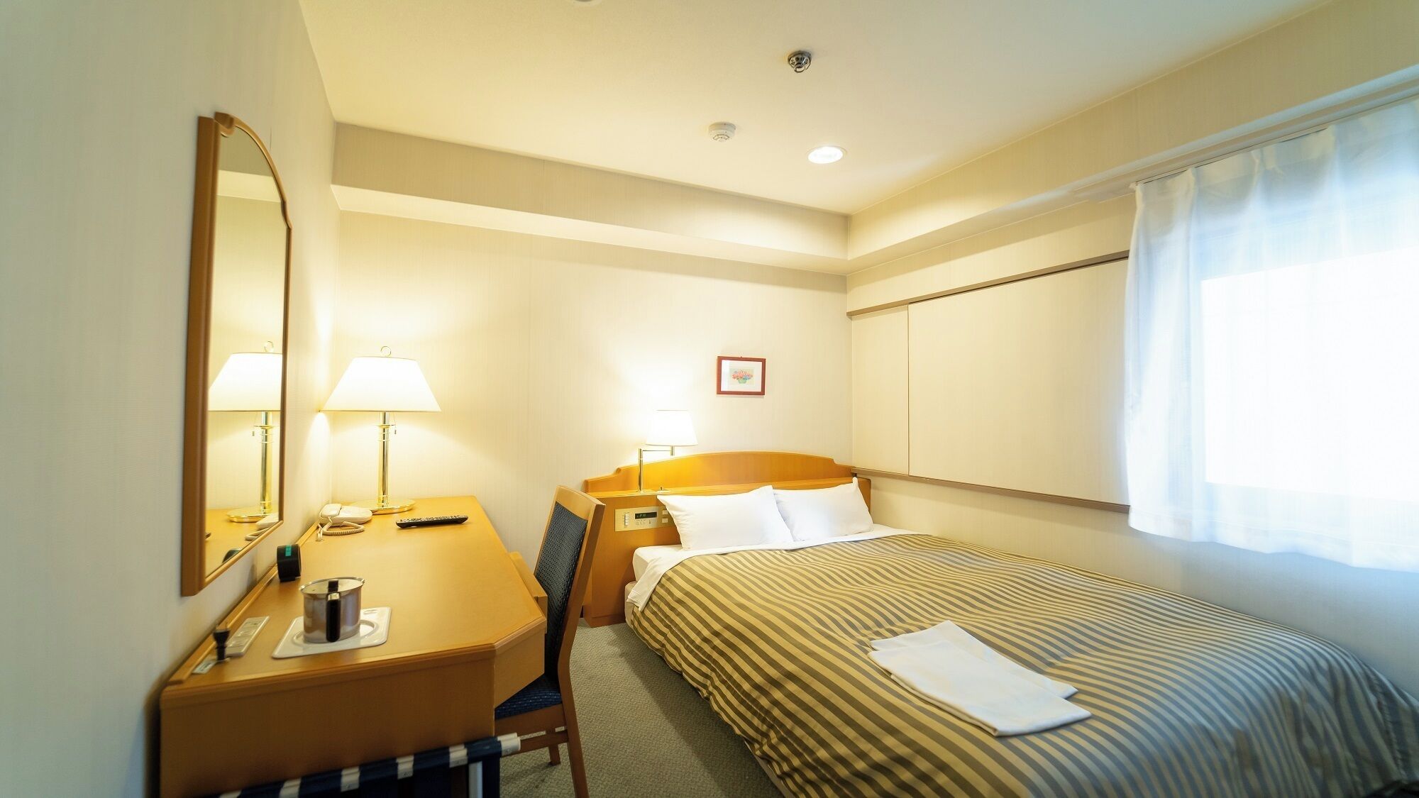 Une chambre d’hôtel avec un lit, un bureau avec une lampe, un téléphone et une petite table avec une tasse et un récipient.