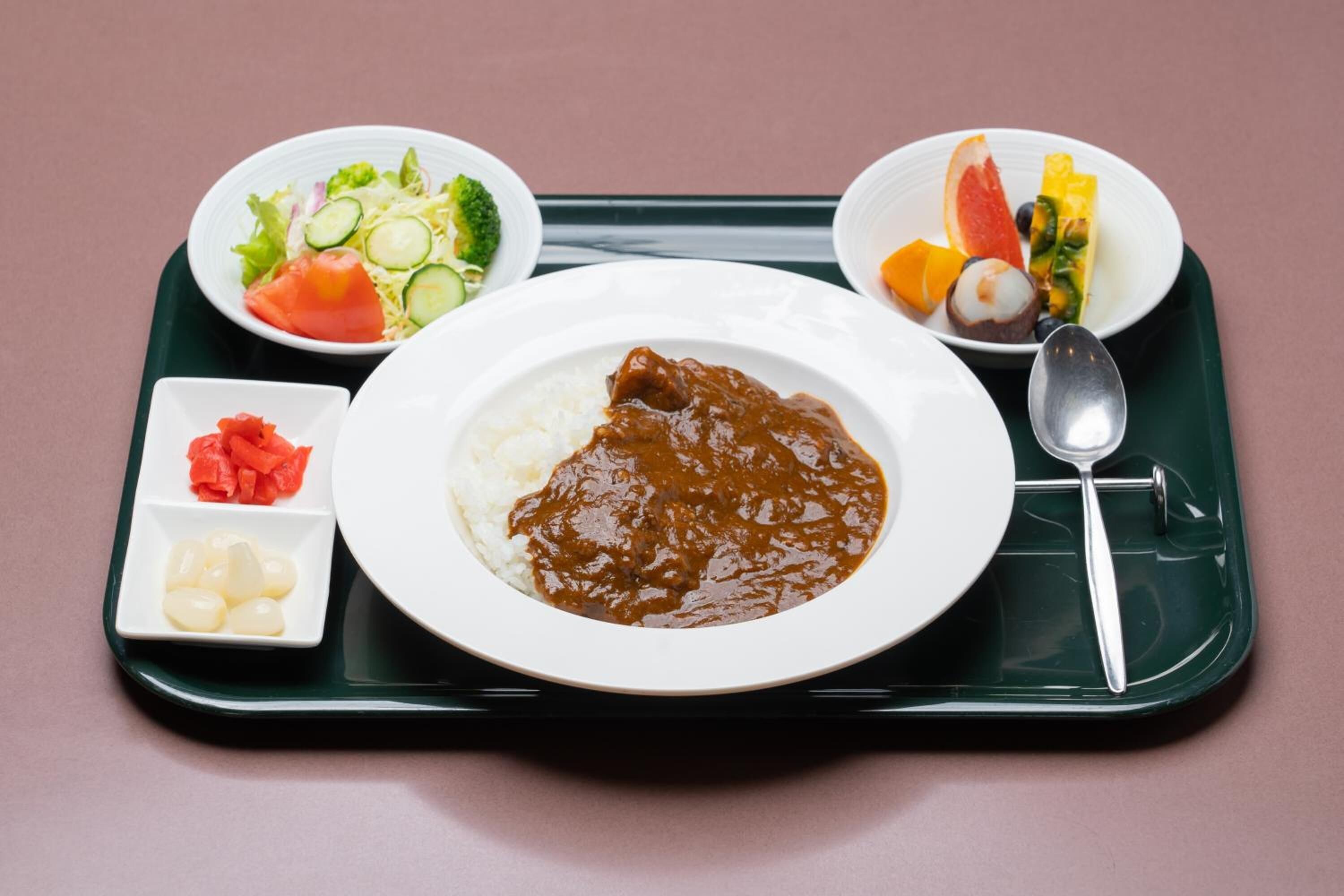 Una bandeja con curry, arroz y verduras, incluyendo una ensalada y un cuenco de frutas.