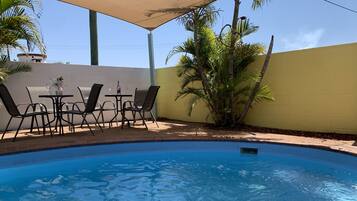 Una piscina con borde de ladrillo, rodeada por un muro amarillo y cubierta por una pérgola.