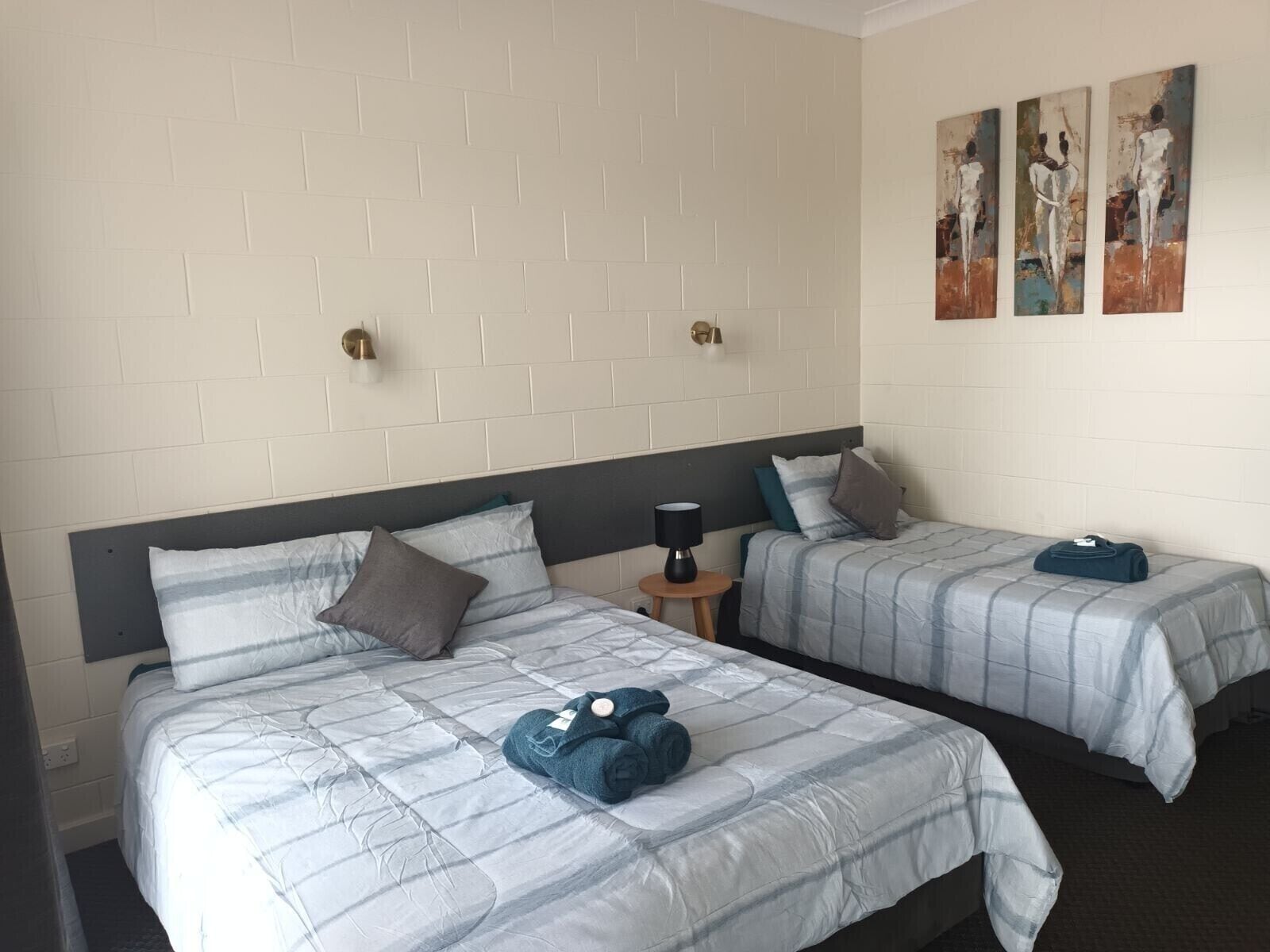 A hotel room with two beds, each with a grey and white striped duvet, a small bedside table with a lamp, and a wall with three abstract paintings.