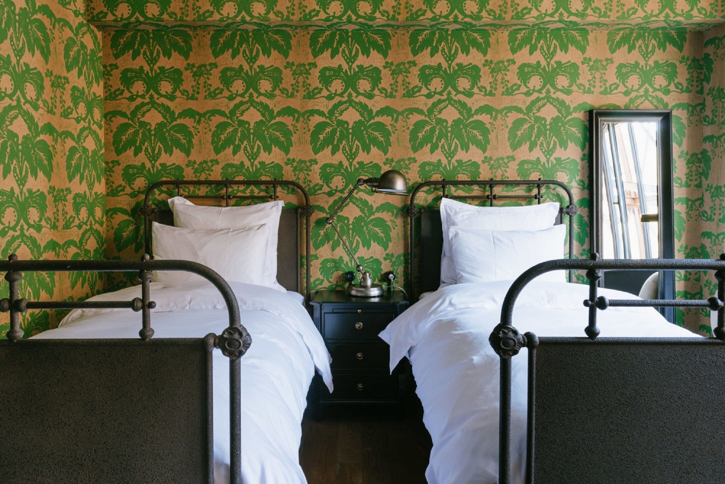 A room with two beds, a nightstand, and a mirror. The walls have a green leafy pattern.
