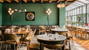 Un restaurant avec des tables rondes et des banquettes, des poutres en bois au plafond et un mur vert orné d’une œuvre d’art circulaire.
