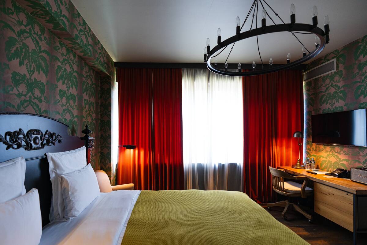 A hotel room with a bed, a desk with a chair, a television, and a chandelier.