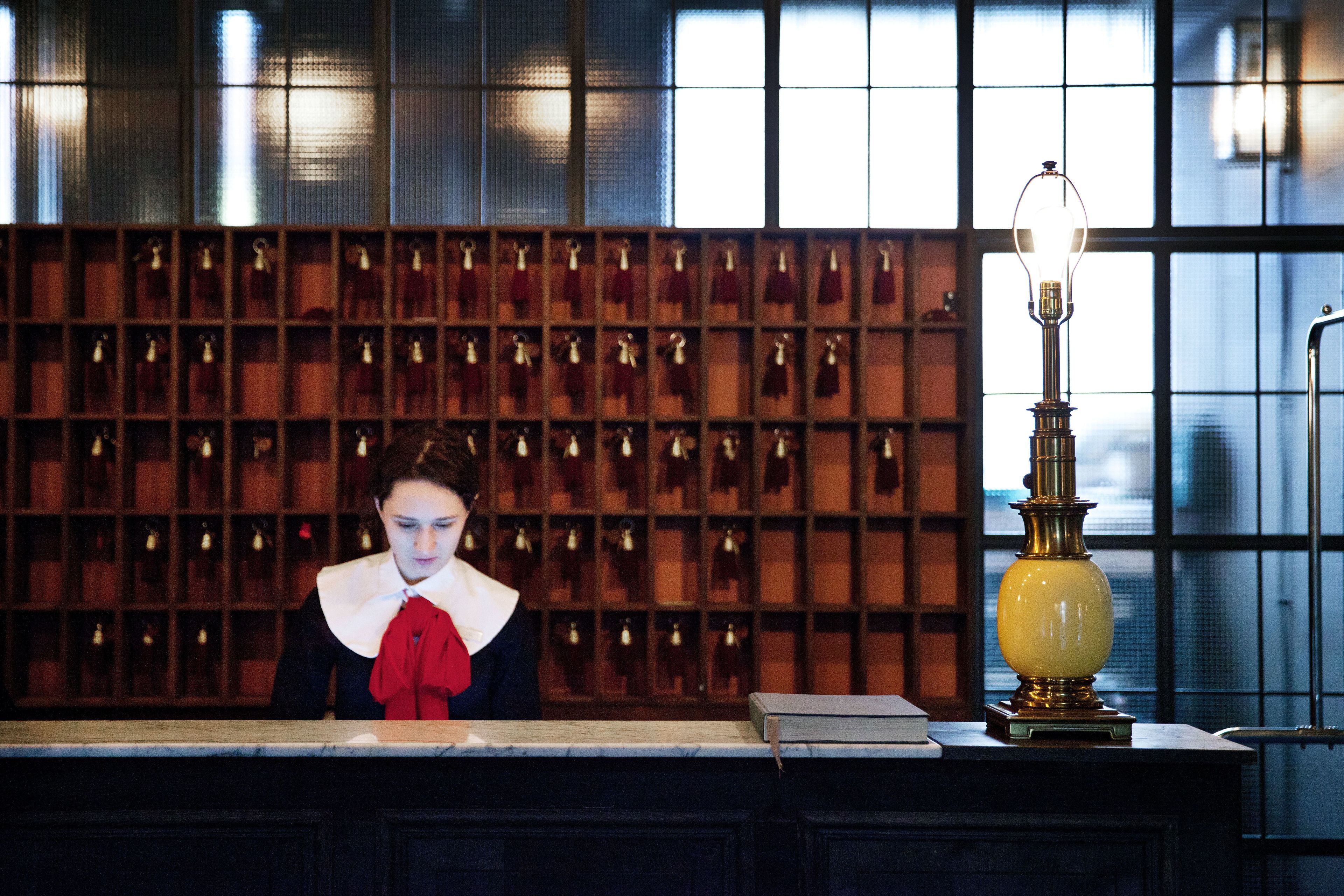 A reception desk with a woman standing behind it, a lamp on the right, and a wall of framed items behind her.